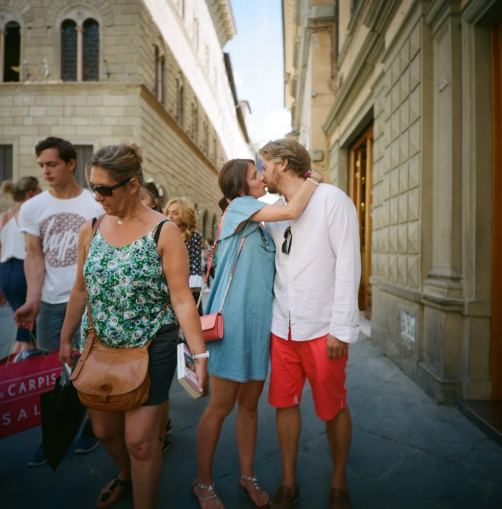 Una coppia si bacia su una strada affollata di una città europea, con edifici storici in pietra sullo sfondo e altre persone che passeggiano, alcune con borse e abiti estivi.