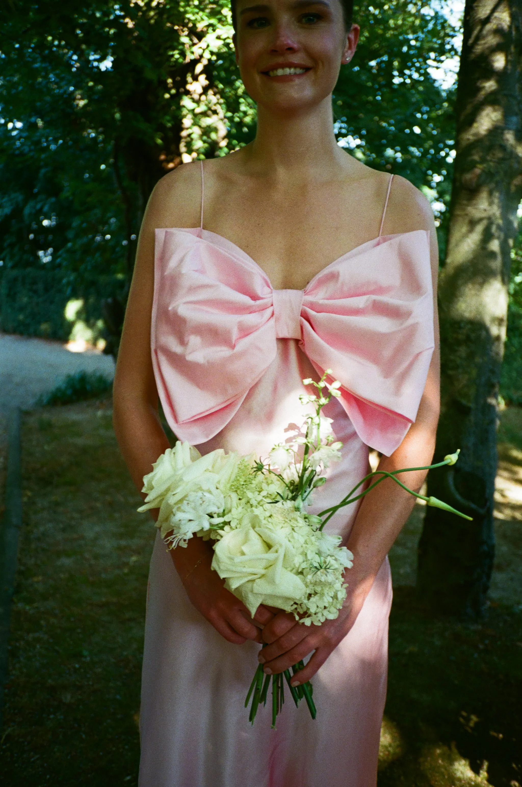 Una donna sorridente indossa un vestito rosa con un grande fiocco sul petto e tiene un bouquet di fiori bianchi, sotto un albero in un ambiente esterno, probabilmente in un parco.