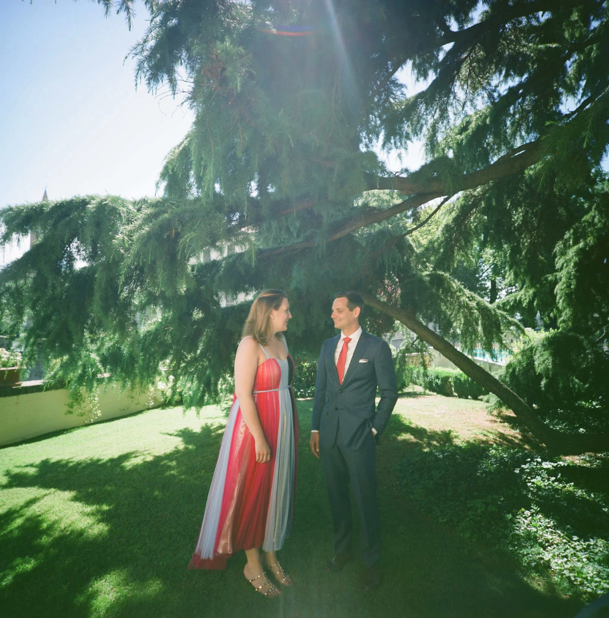 Uomo e donna si tengono per mano sotto un albero, si sorridono e indossano abiti eleganti in un parco soleggiato.