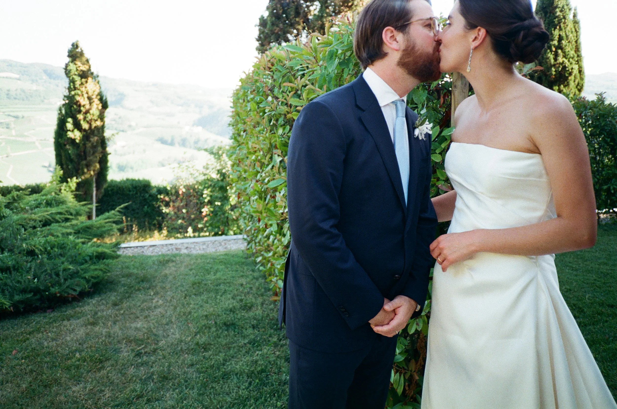 Un uomo e una donna si baciano durante un matrimonio all'aperto, con un paesaggio collinare verde sullo sfondo.