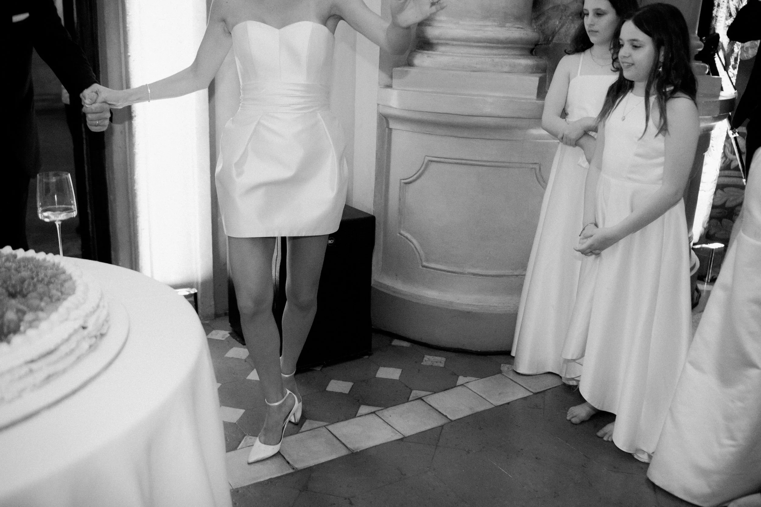 Immagine in bianco e nero di una scena di un matrimonio con persone in abiti formali, tra cui una donna con un vestito bianco che pare far parte di una cerimonia.