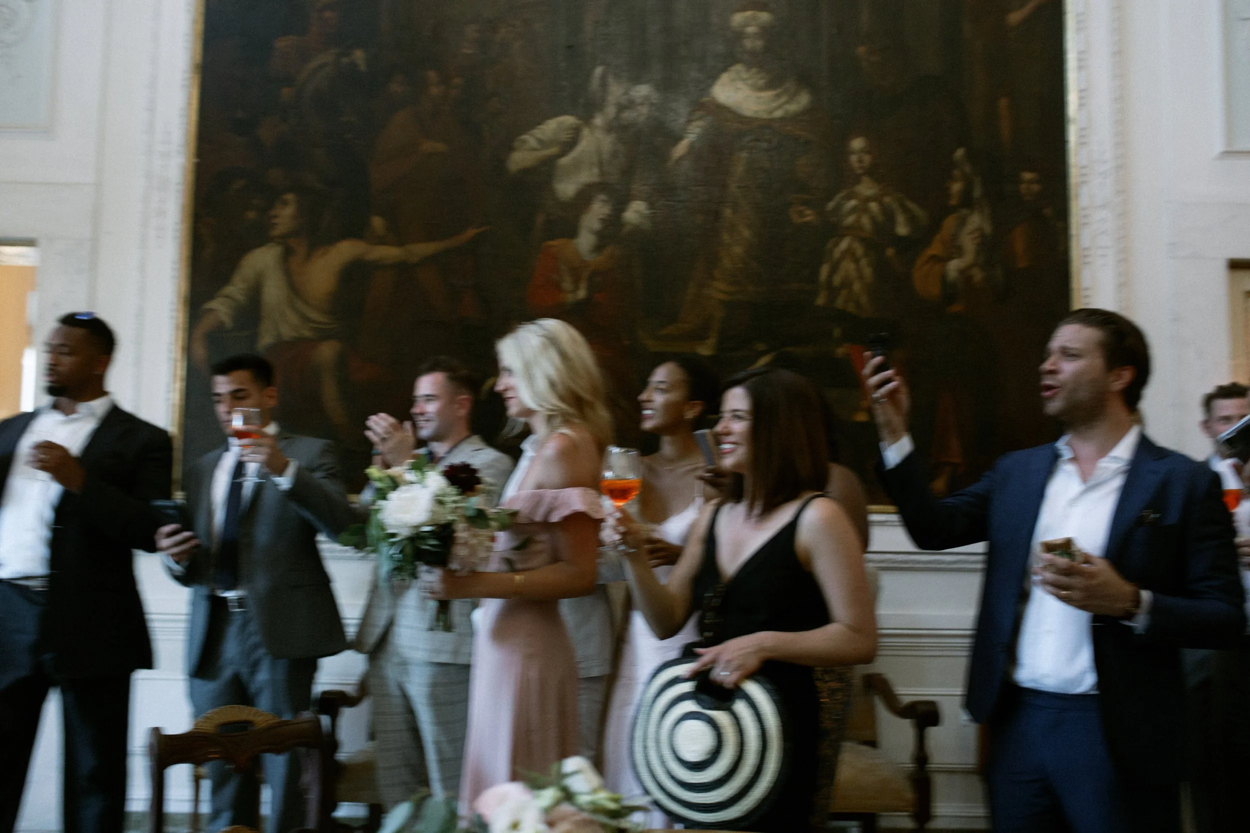 Un gruppo di persone eleganti che partecipano a una festa o un evento, con alcuni che brindano e sorridono, in un ambiente raffinato con un grande quadro storico sul muro.