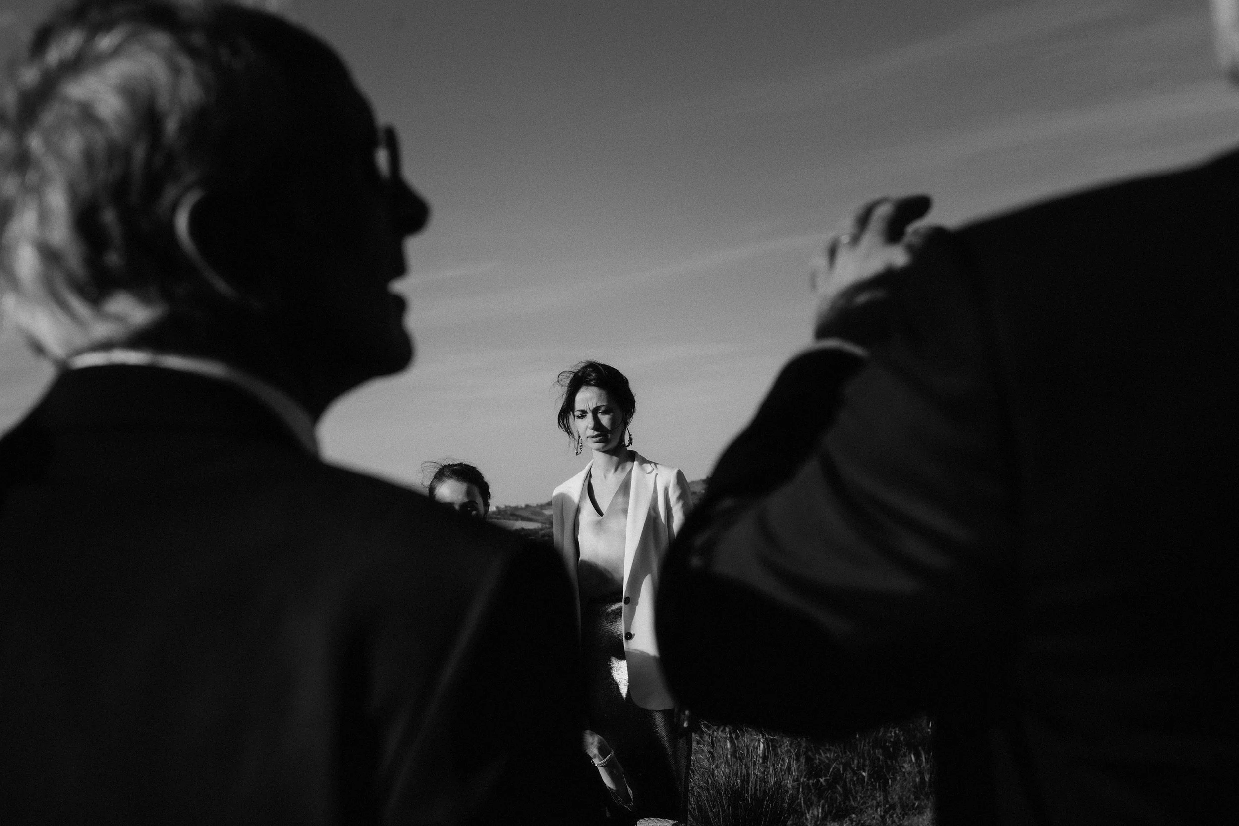 Una scena in bianco e nero di tre persone all'aperto, con un uomo con i capelli bianchi e una donna con i capelli scuri in primo piano, mentre un'altra donna con abiti chiari guarda verso i due e osserva attentamente. La scena ha un'atmosfera seria e contemplativa.