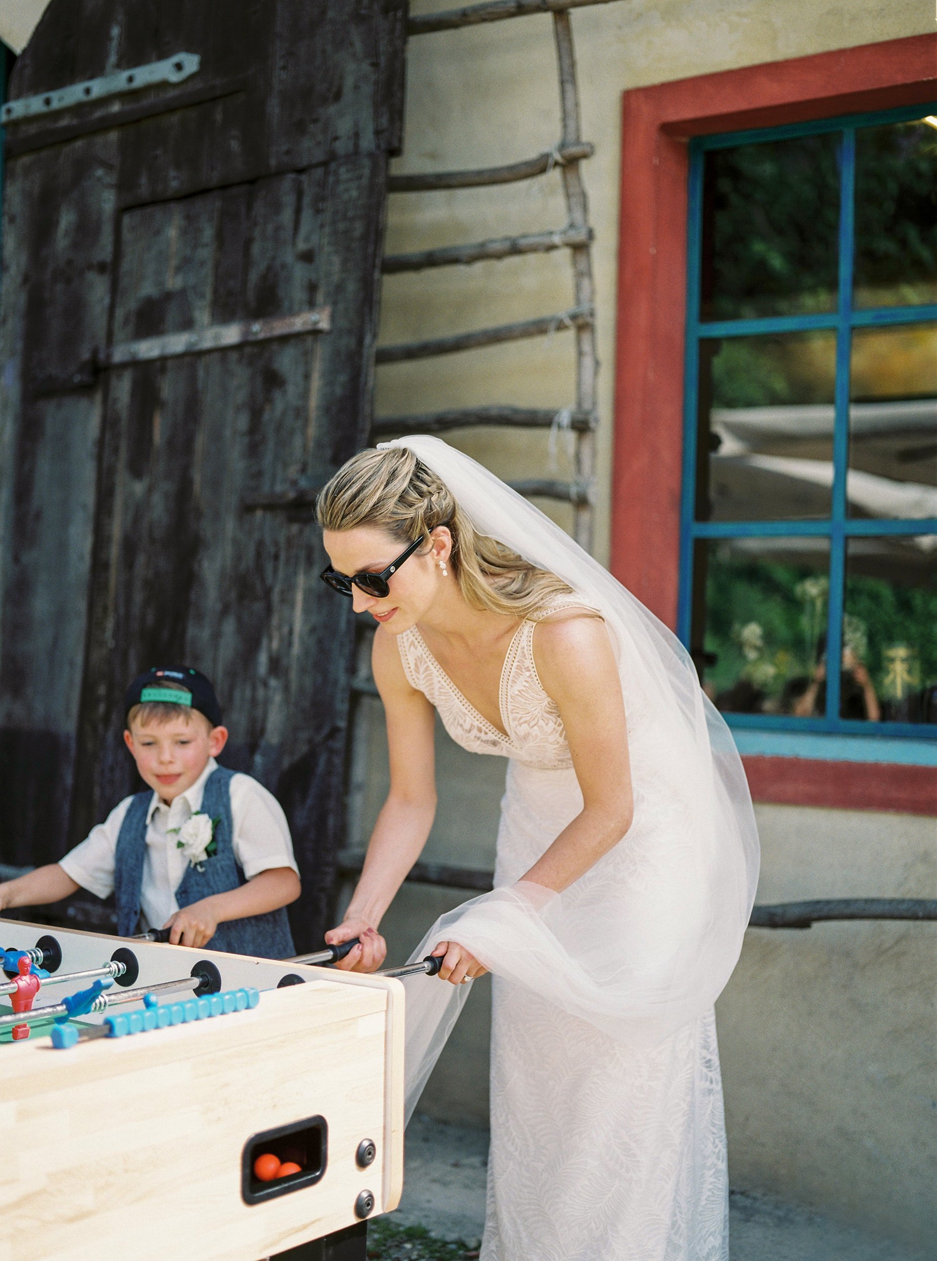 Una donna in abito da sposa con capelli raccolti e occhiali da sole sta giocando a calcio balilla con un bambino che indossa un cappellino, davanti a una parete con finestra blu. Sono all'aperto, in un ambiente rustico.