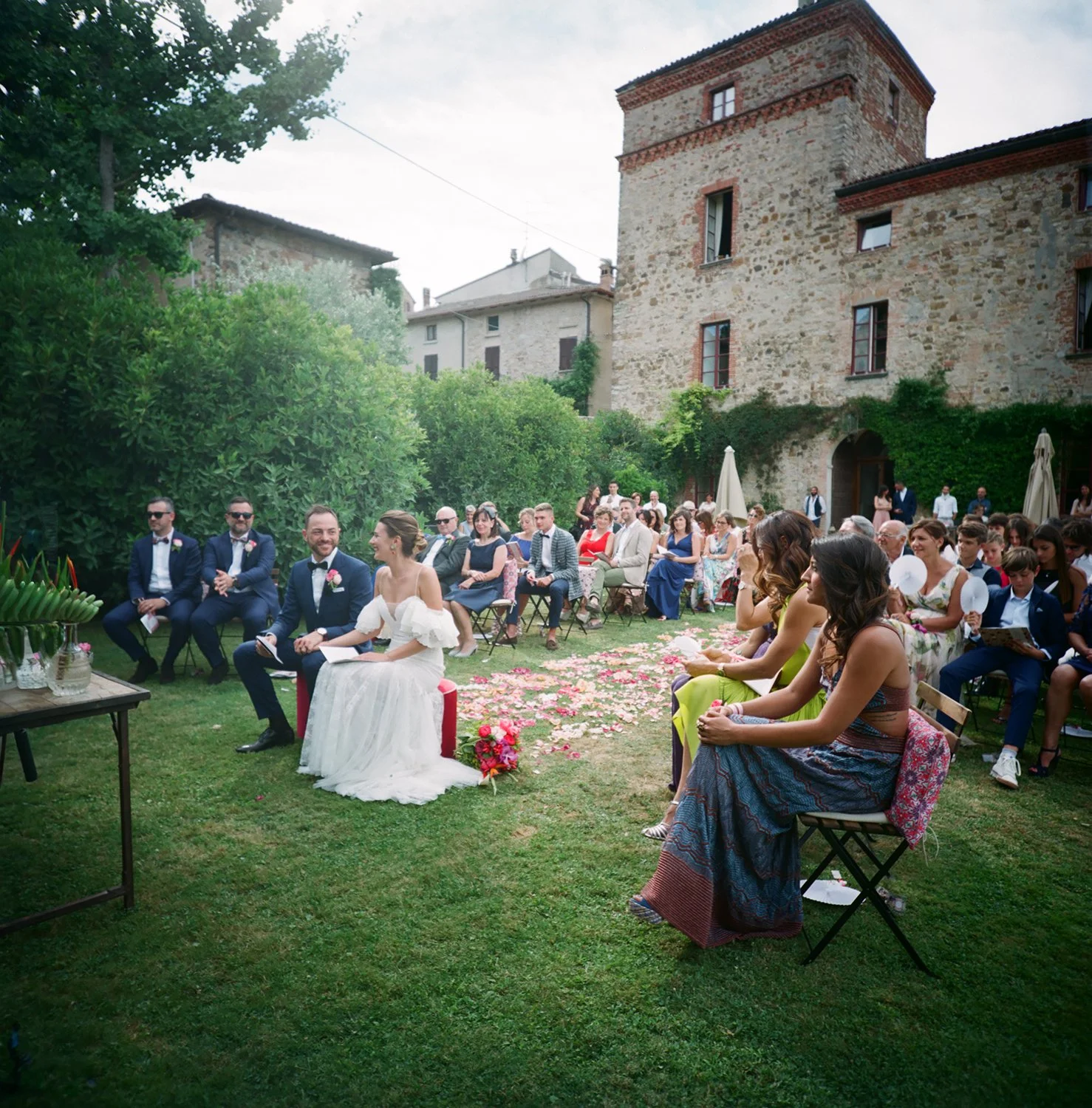 Cerimonia di matrimonio all'aperto con sposi seduti davanti a un pubblico seduto su sedie in un giardino. Sullo sfondo si vede un vecchio edificio in pietra.
