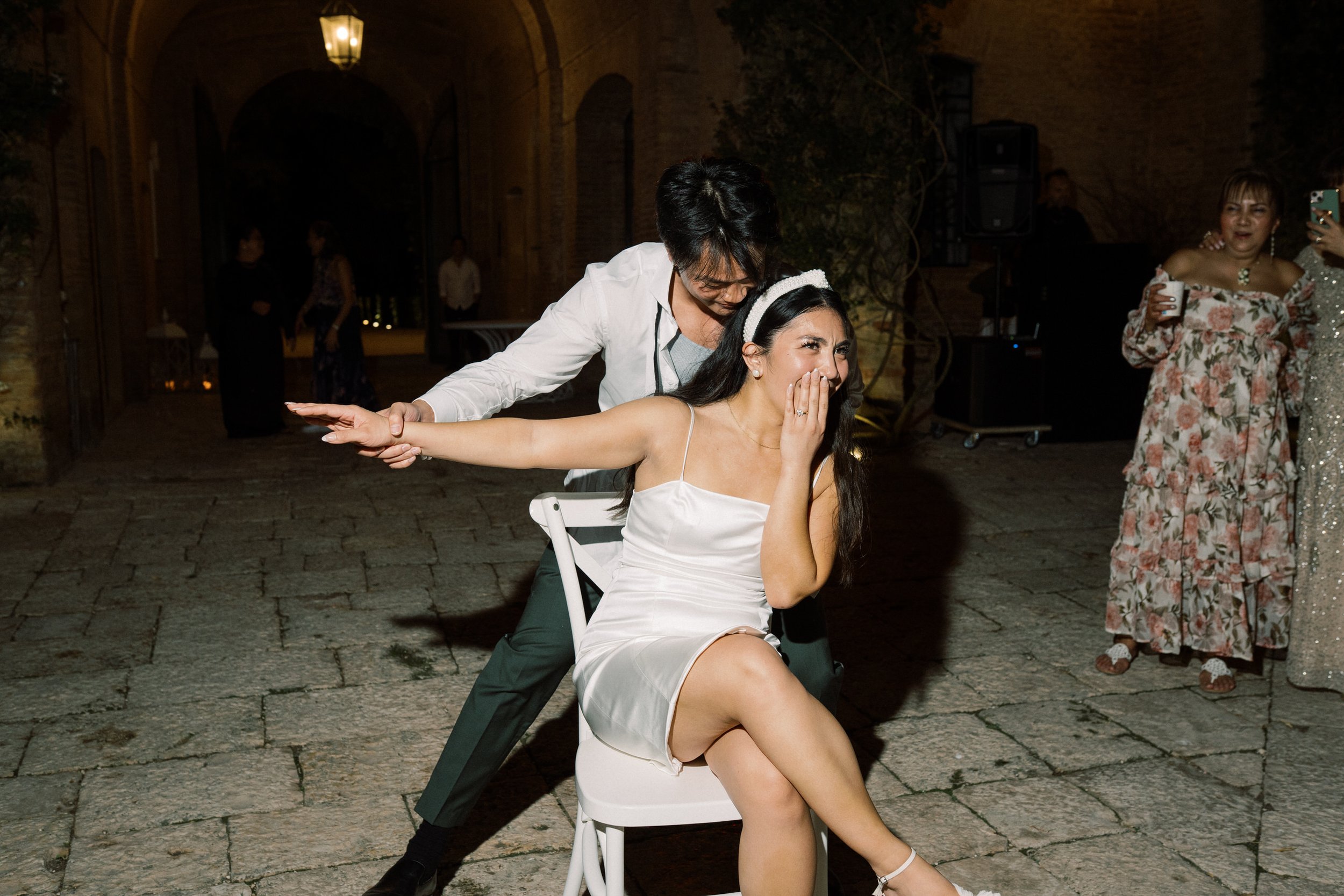 Una donna in abito da sposa ride coprendosi la bocca, seduta su una sedia, mentre un uomo si avvicina da dietro e la prende in braccio in un momento di festa.