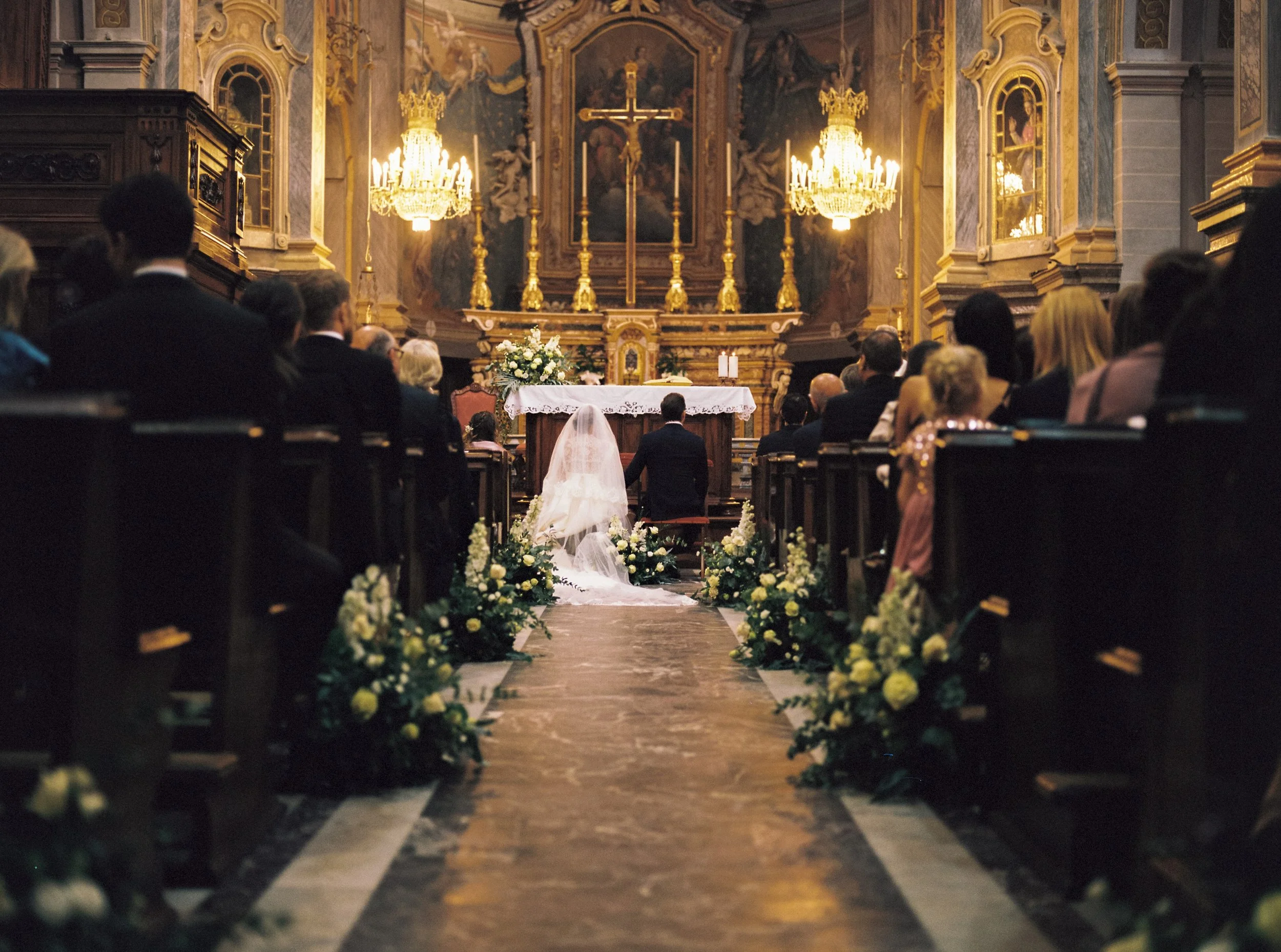 Interno di una chiesa decorata con candelabri dorati e un altare con un crocifisso, presente una folla di persone riunite per una cerimonia, probabilmente un matrimonio.