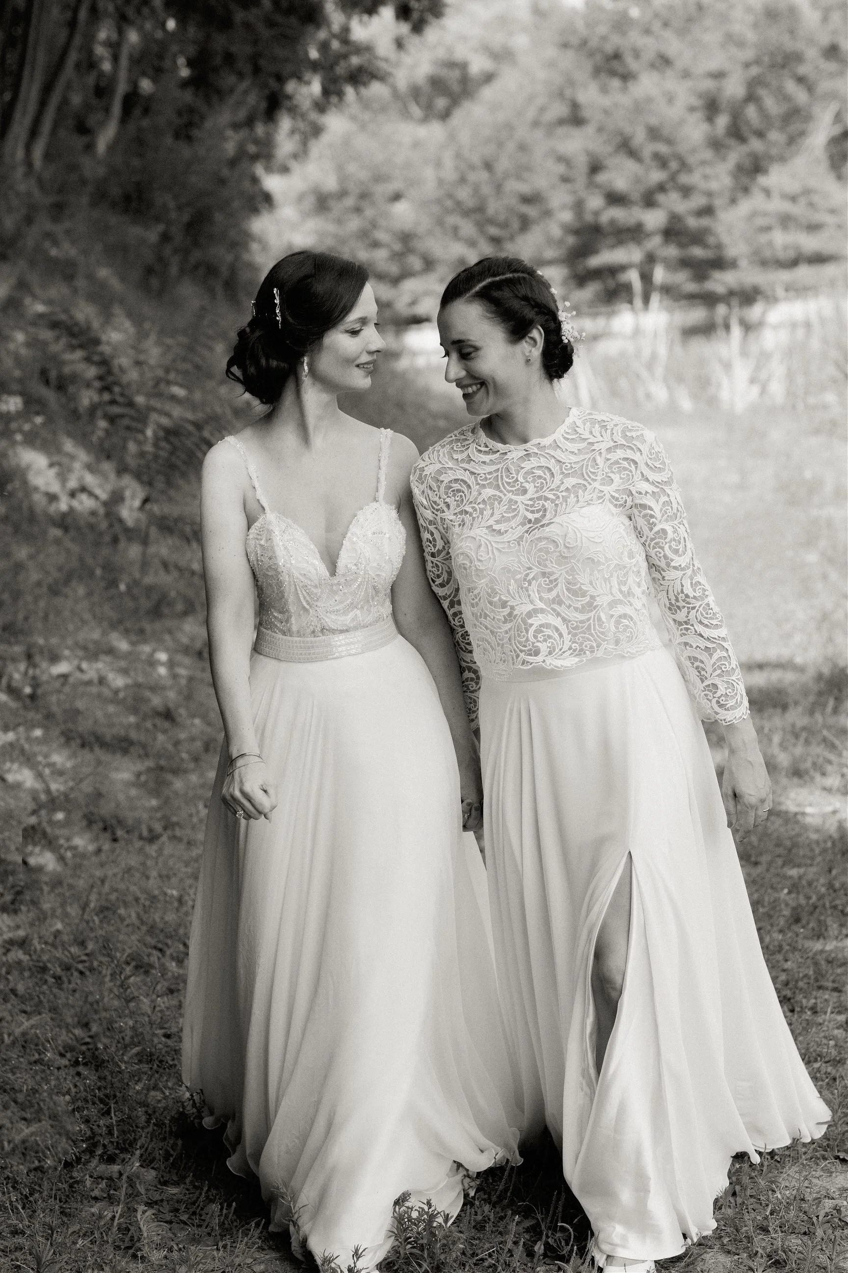 Due donne in abiti da sposa che tengono le mani e si sorridono, ambientate all'aperto