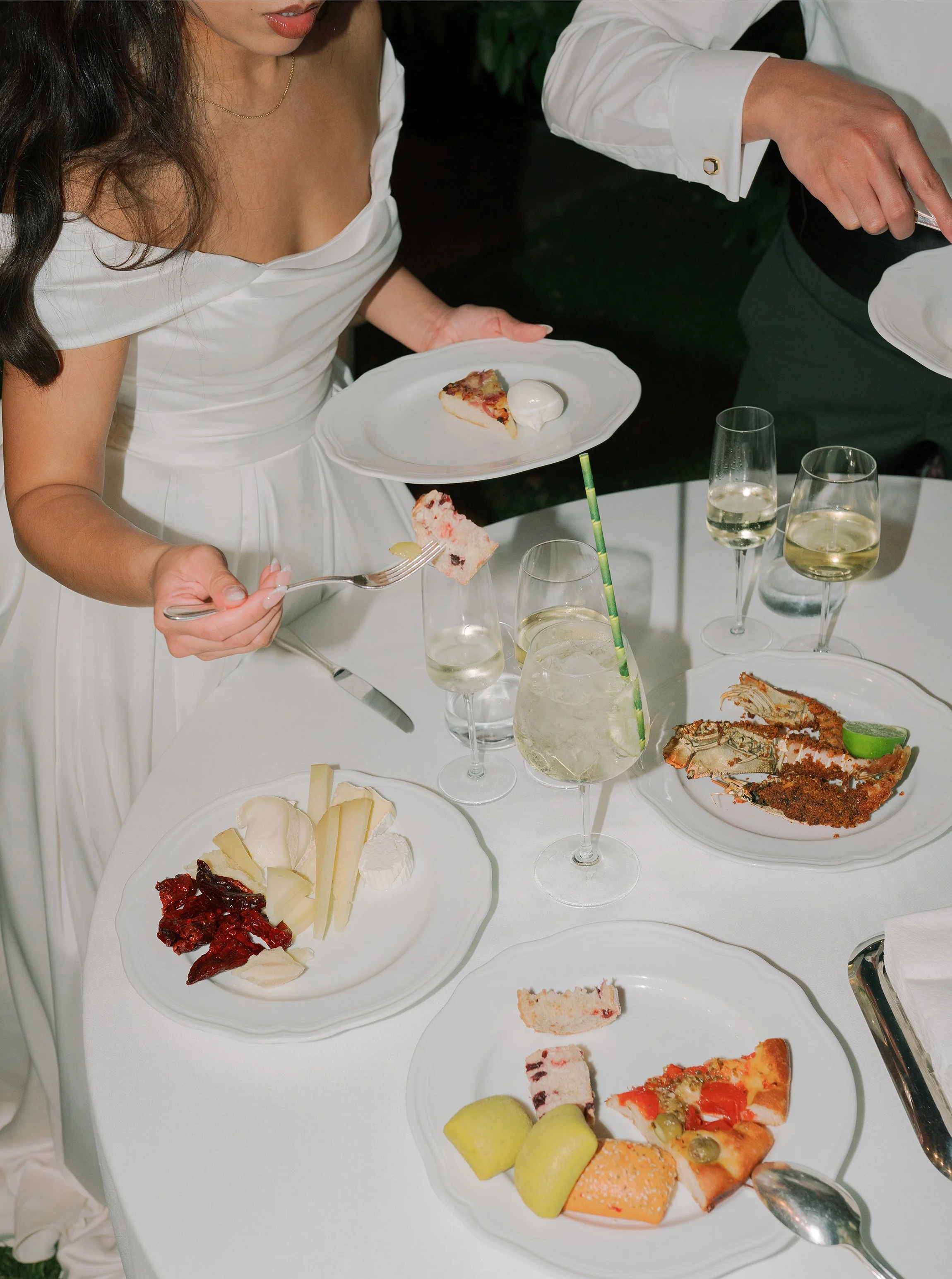 Donna con vestito bianco che tiene un piatto con una fetta di pizza e un palloncino decorativo verde e bianco alla festa. Tavola apparecchiata con vari piatti di formaggi, dolci e snack, bicchieri di vino bianco, e un drink con limone e cannucce verdi ai piedi della donna.