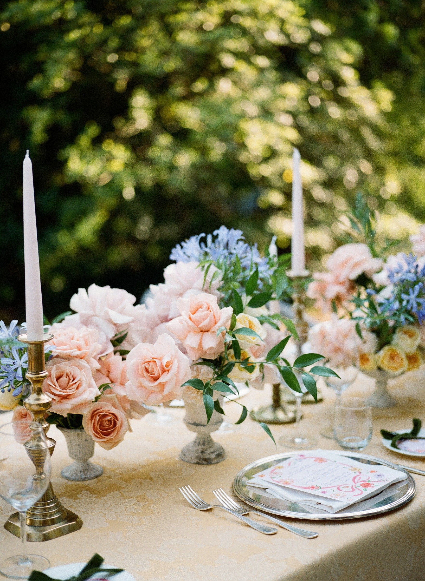 Tavola di matrimonio decorata con fiori rosa e bianchi, candelabri e tovaglioli con menu floral, sotto un grande albero con luce solare filtrata