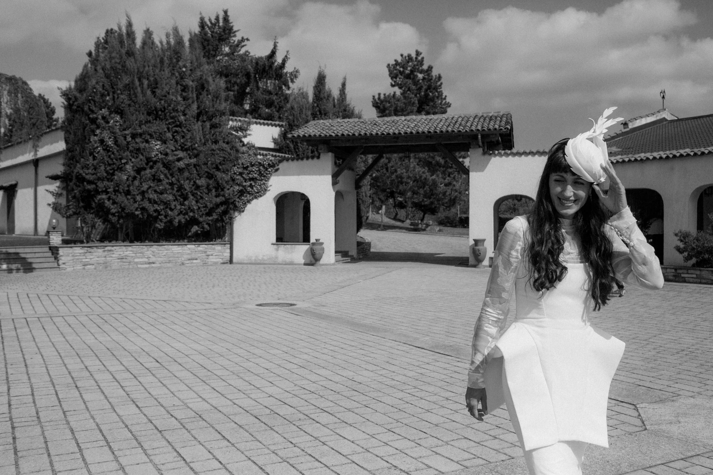 Una donna con abito bianco e cappello bianco con decorazione scherma una risata mentre si sistema il cappello, in un cortile con archi e piante.