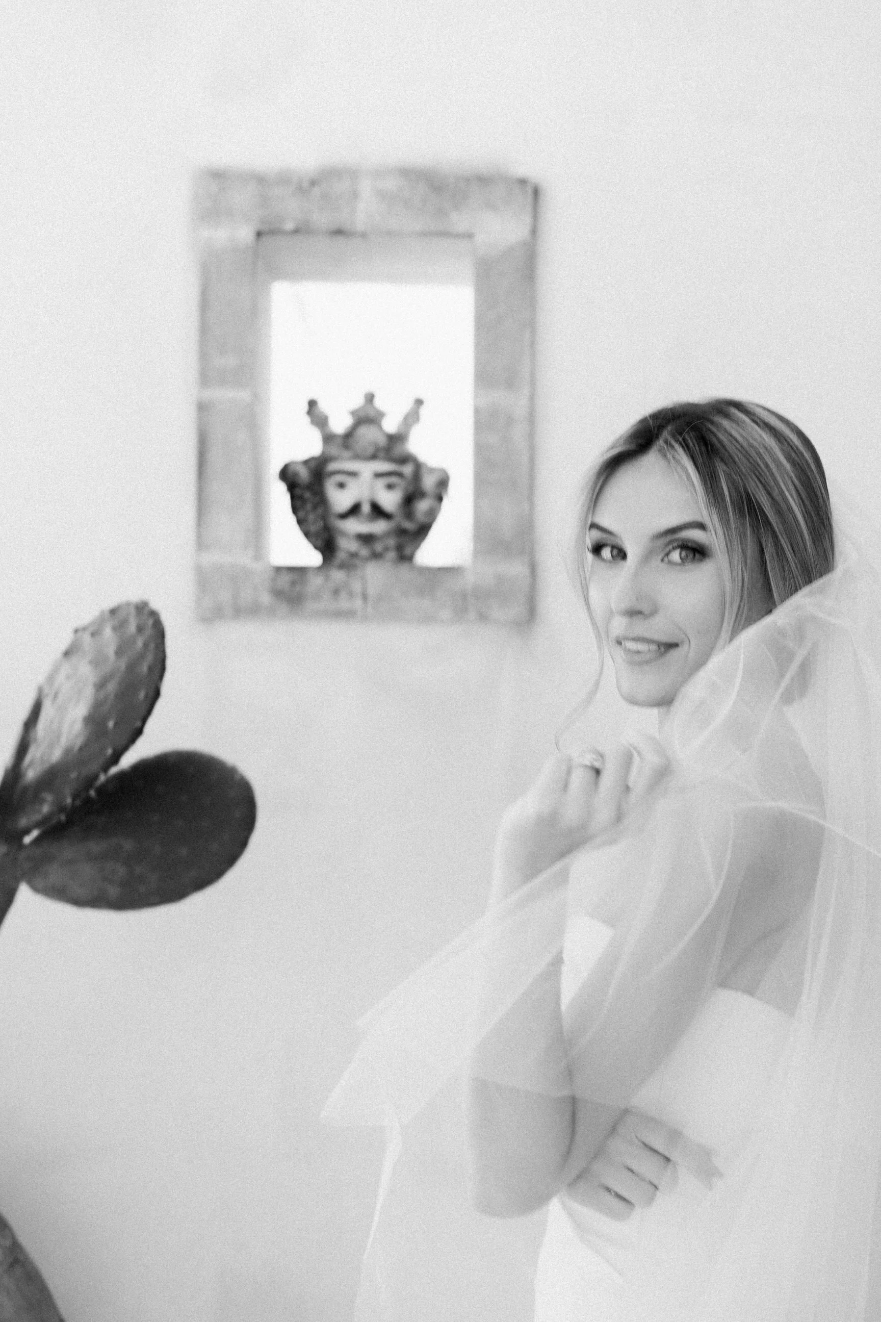 Ragazza con capelli lisci e occhi azzurri, in un'ambientazione bianca, volto e spalle visibili, con una mano vicino al viso. Sullo sfondo, un volto di una testa con corona decorativa e un quadro con un motivo architettonico.