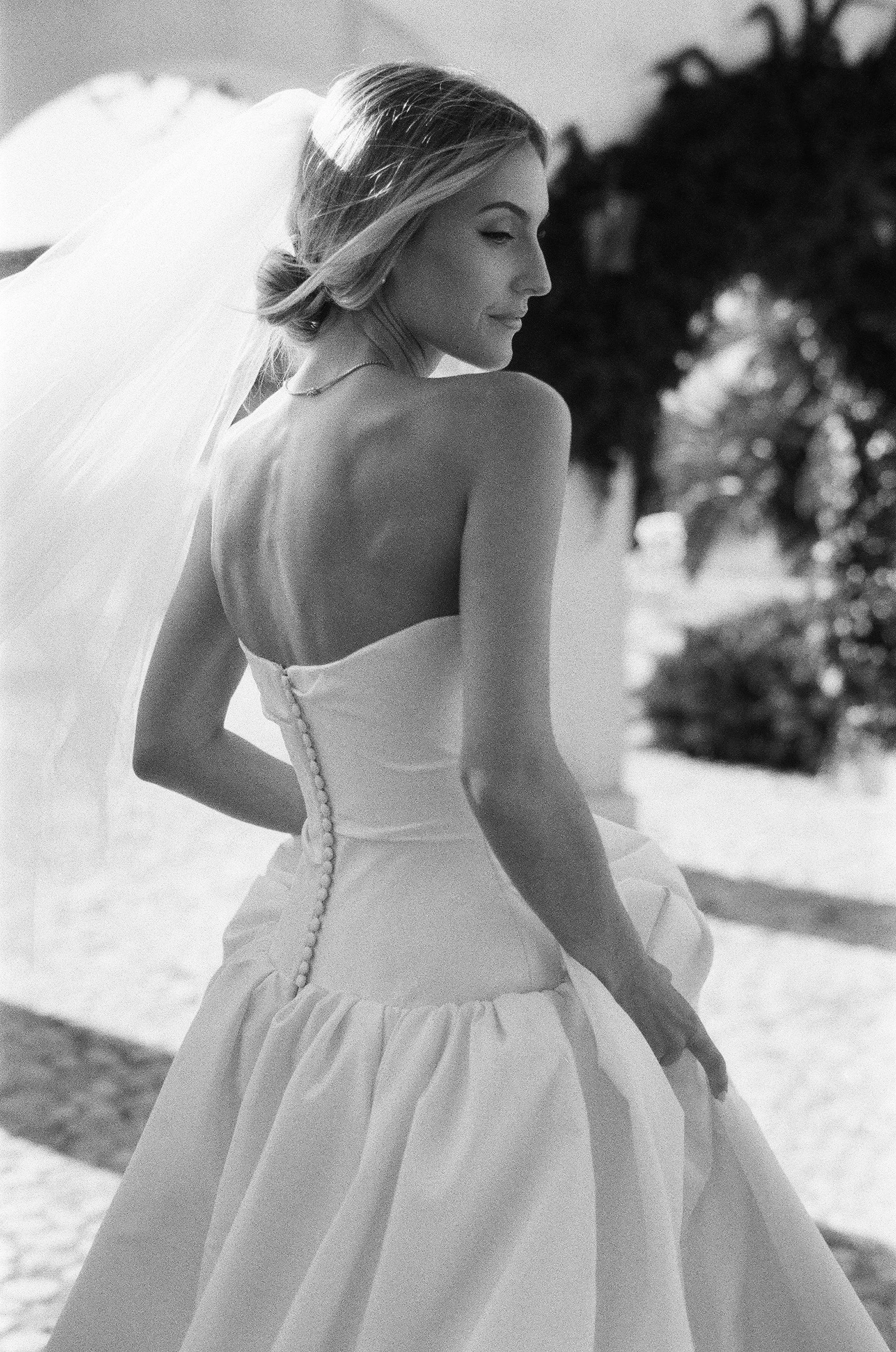 Ritratto di una donna in abito bianco, vista di profilo, con capello arricciato, sullo sfondo di alberi e cielo, in bianco e nero.