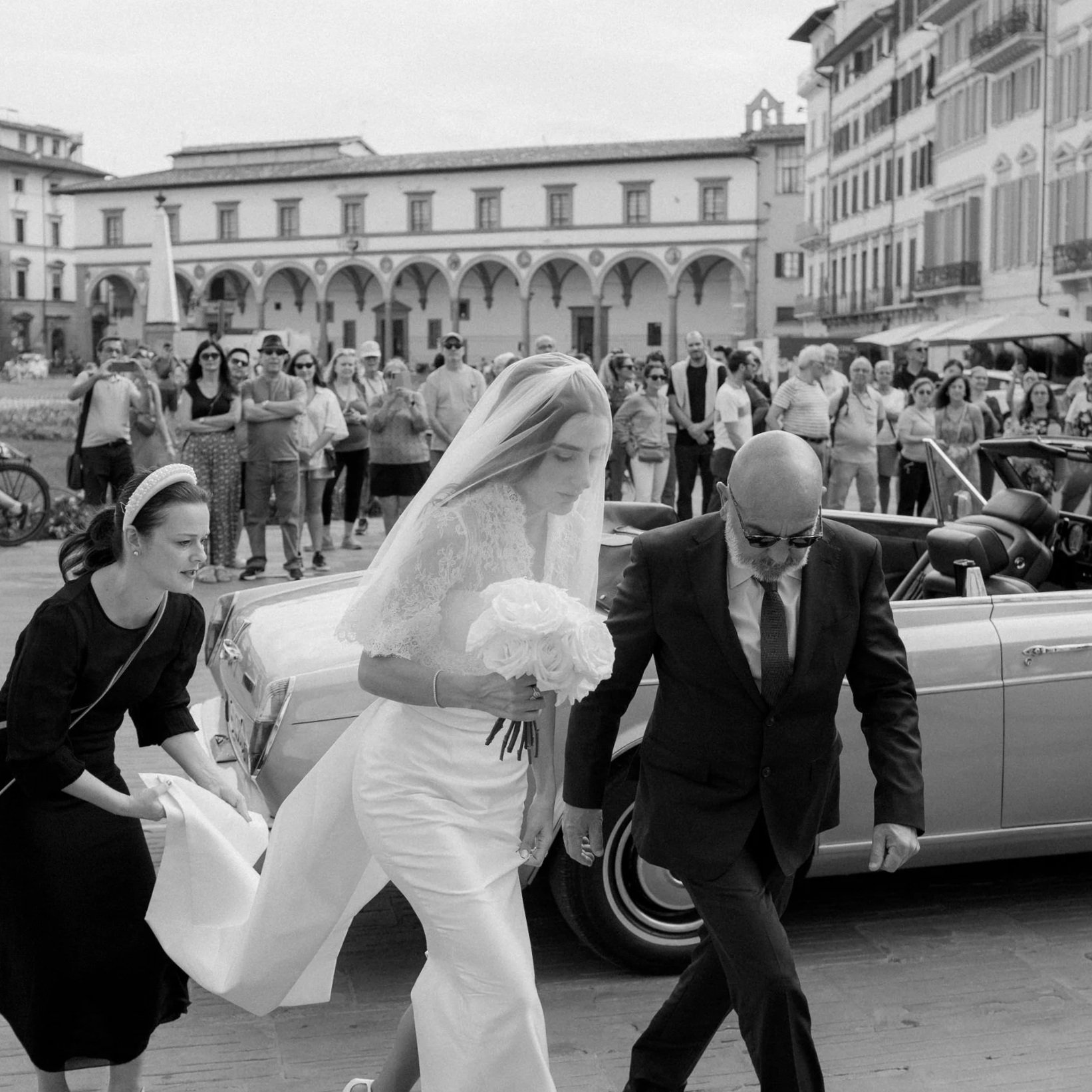 Una donna con abito bianco e velo tiene un mazzo di fiori mentre scende da un'auto d'epoca in una piazza affollata, accompagnata da un uomo con abito scuro. Sullo sfondo si vedono molte persone e edifici storici.