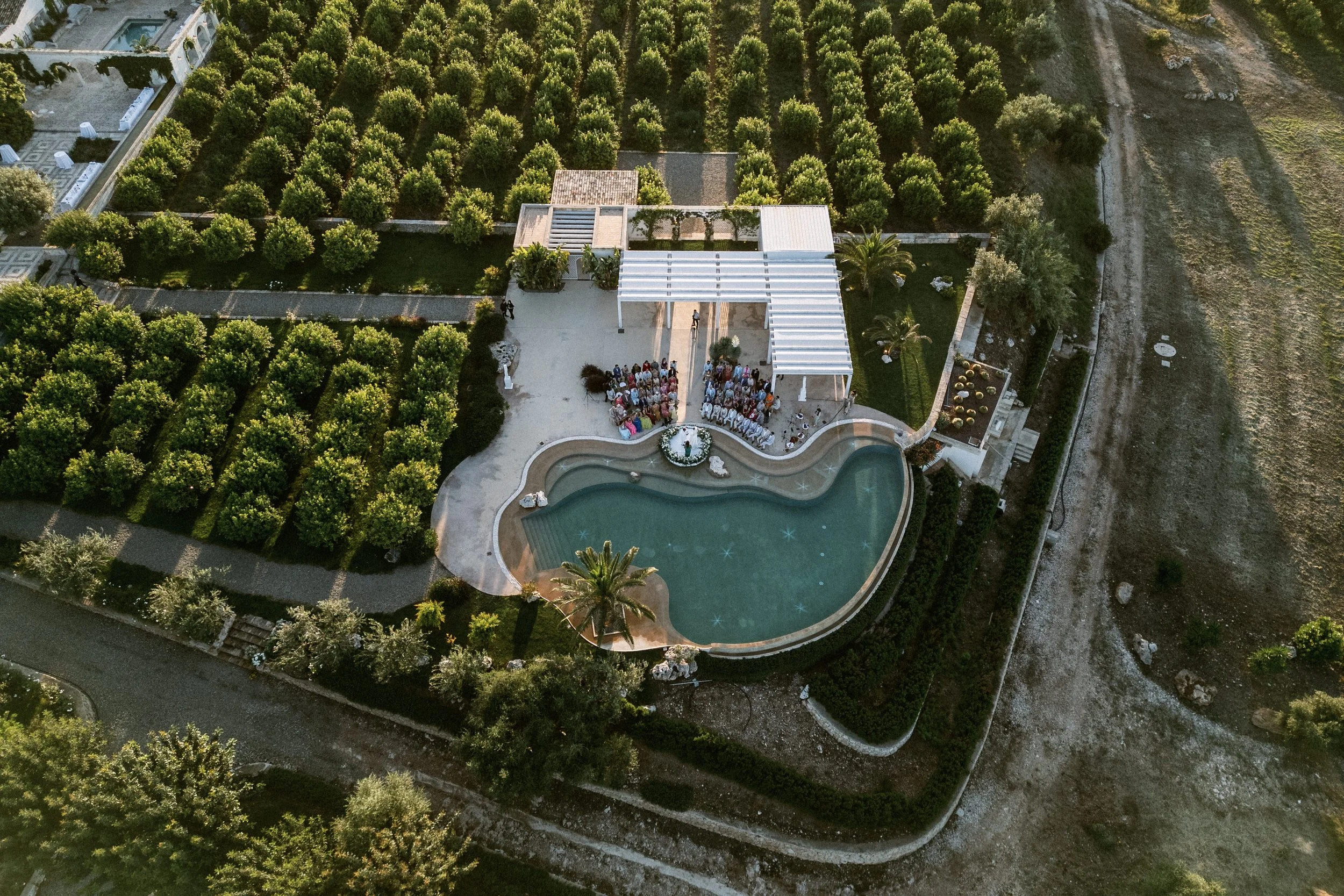Vista aerea di un giardino con piscina, sedie e un gruppo di persone riunite, circondato da alberi e vegetazione, con una struttura coperta e un sentiero.