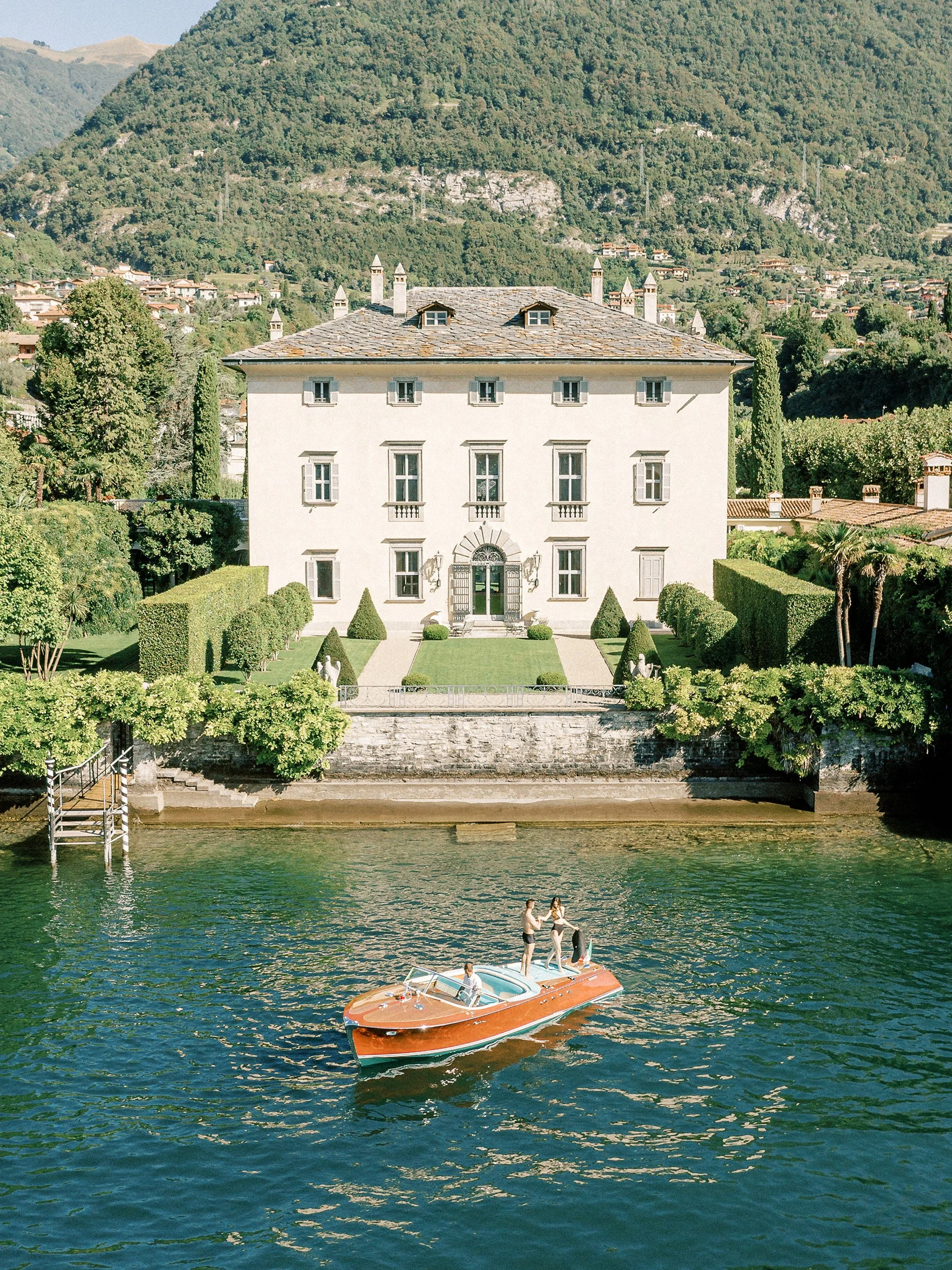 Una barca a motore rossa con persone sul lago in fronte a una villa imponente con giardino e montagne sullo sfondo.