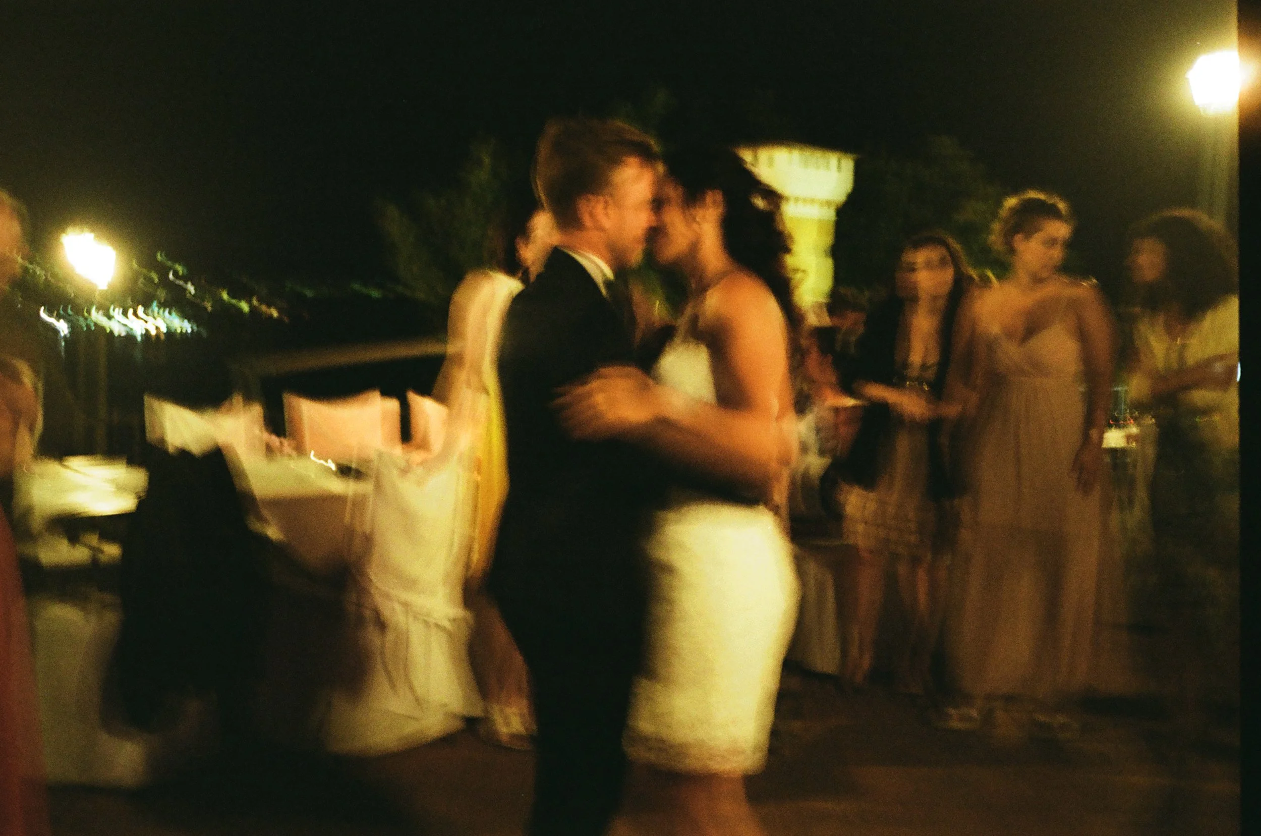 Persone che si abbracciano in un matrimonio, in serata all'aperto con luci calde e sfondo scuro.
