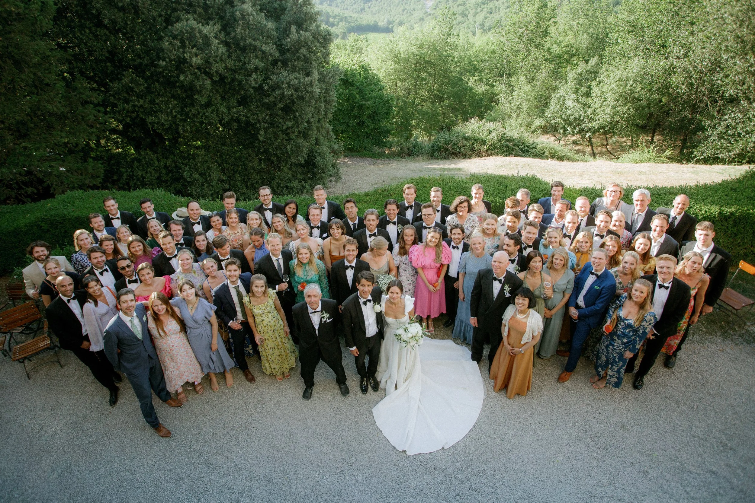Gruppo di persone in abiti eleganti e casual sotto gli alberi in un parco, sembrano partecipare a un matrimonio o un evento formale, con molte persone sorridenti e festose.