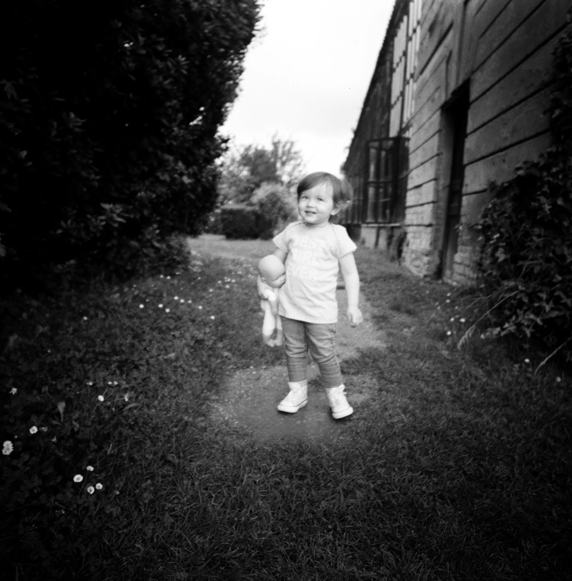 Bambina sorridente che cammina lungo un sentiero all'aperto, circondata da cespugli e vicino a una casa in muratura, in bianco e nero.