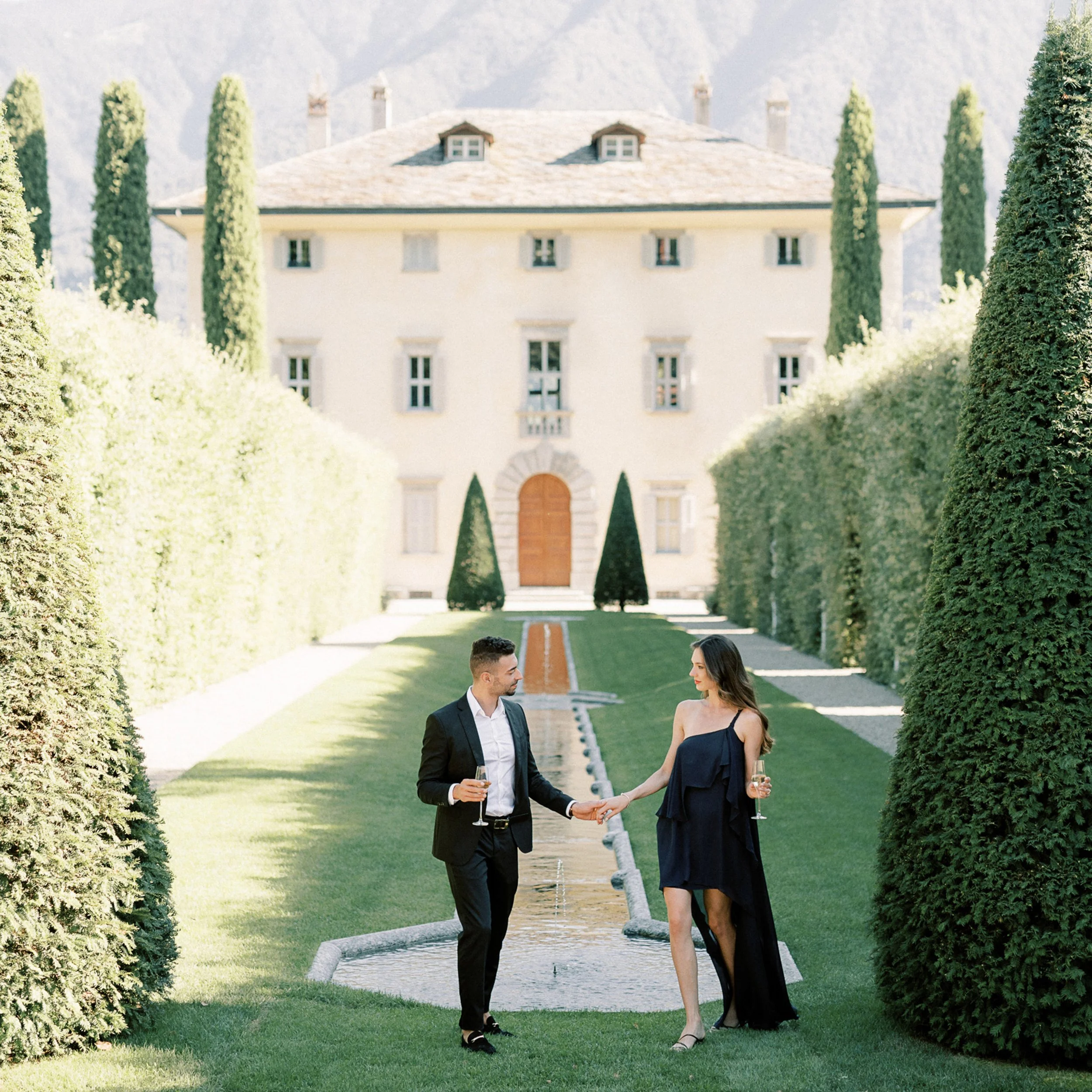 Coppia che si tiene per mano con bicchieri di champagne in un giardino con fontana e un grande edificio sullo sfondo, durante un evento elegante.
