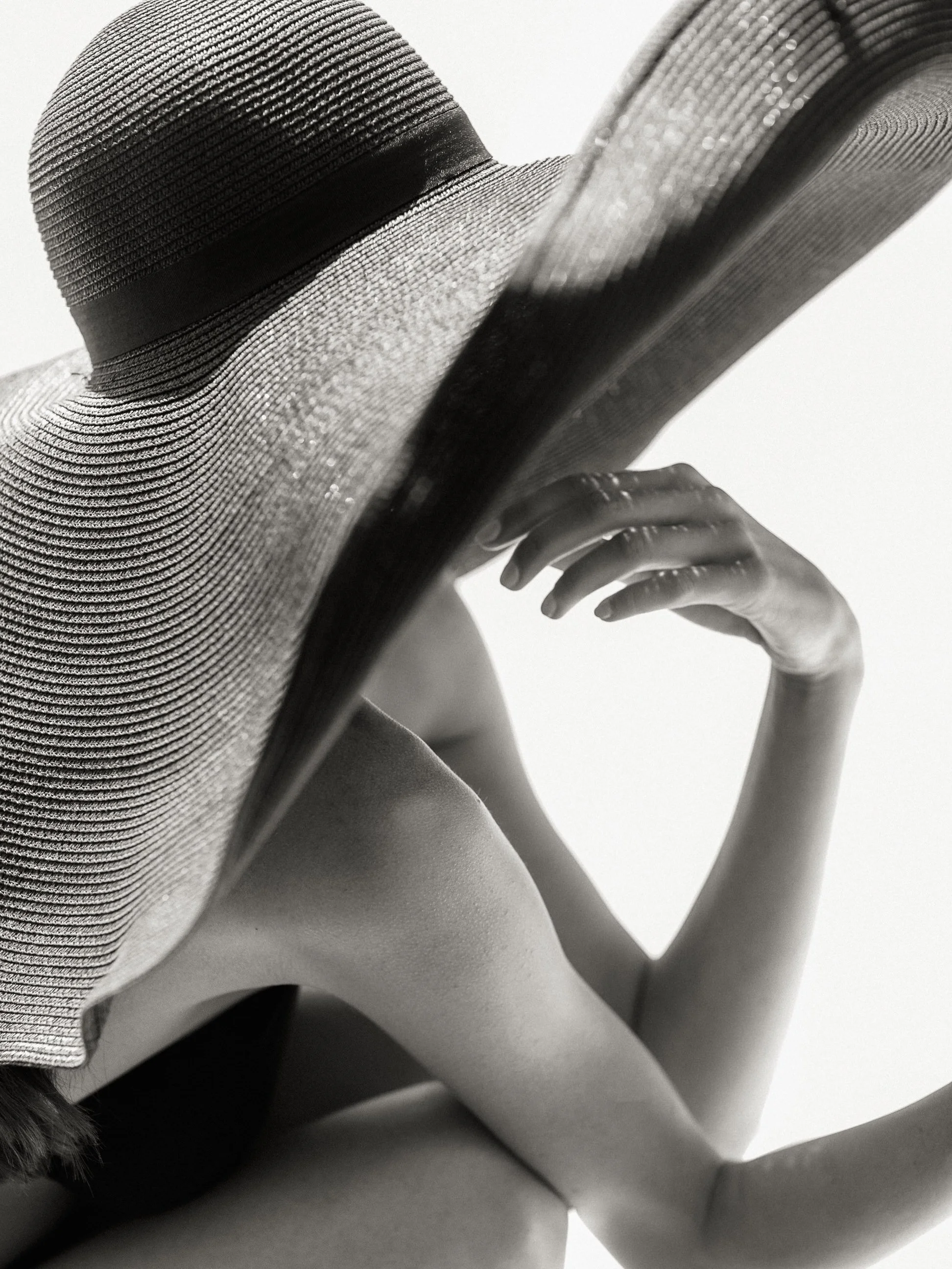 Un cappello di paglia con nastro nero indossato da una figura femminile, vista in prospettiva da un'angolazione ravvicinata in nero e bianco.