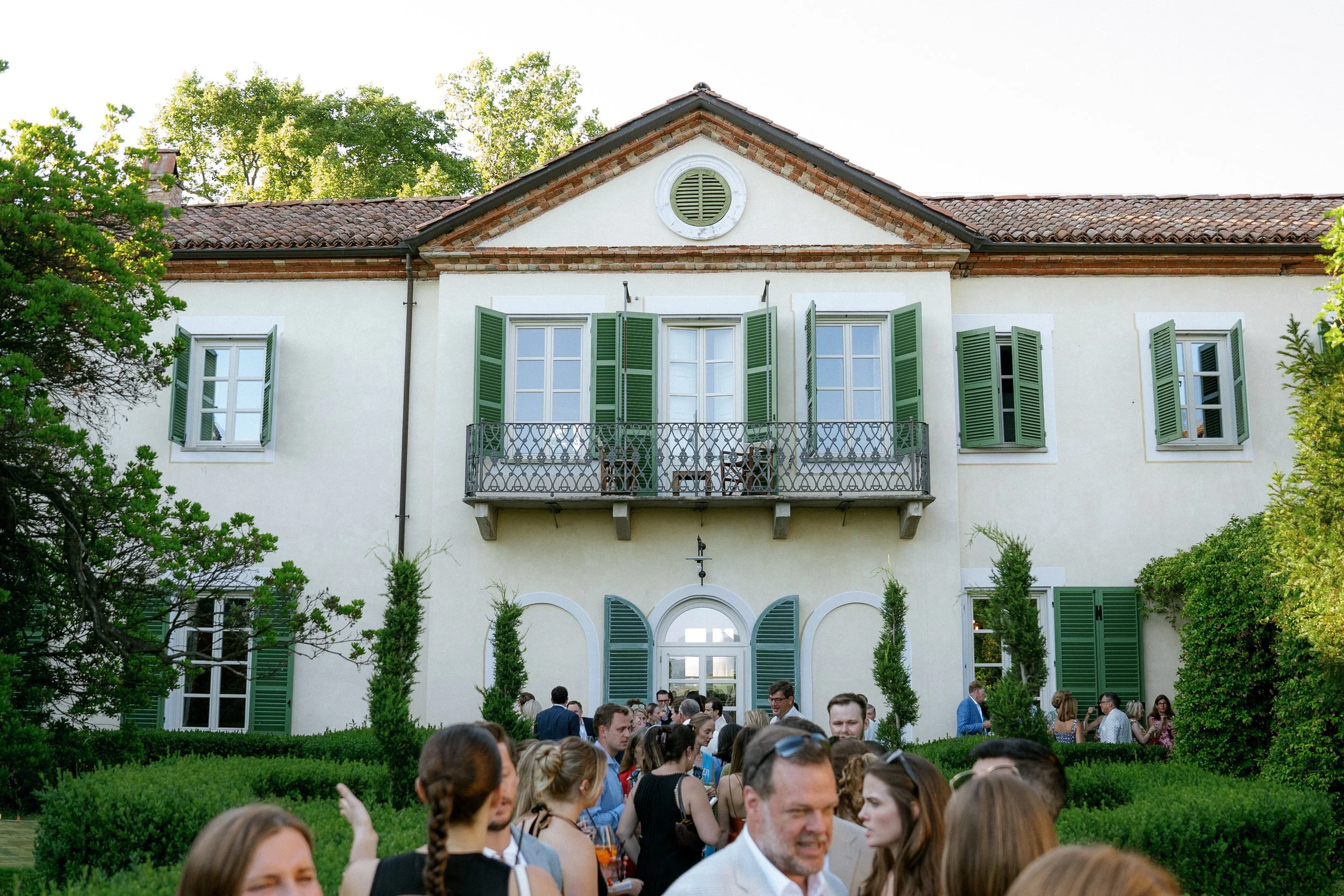 Gruppo di persone che si riuniscono in un giardino davanti a una casa con finestre con persiane verdi e balcone.