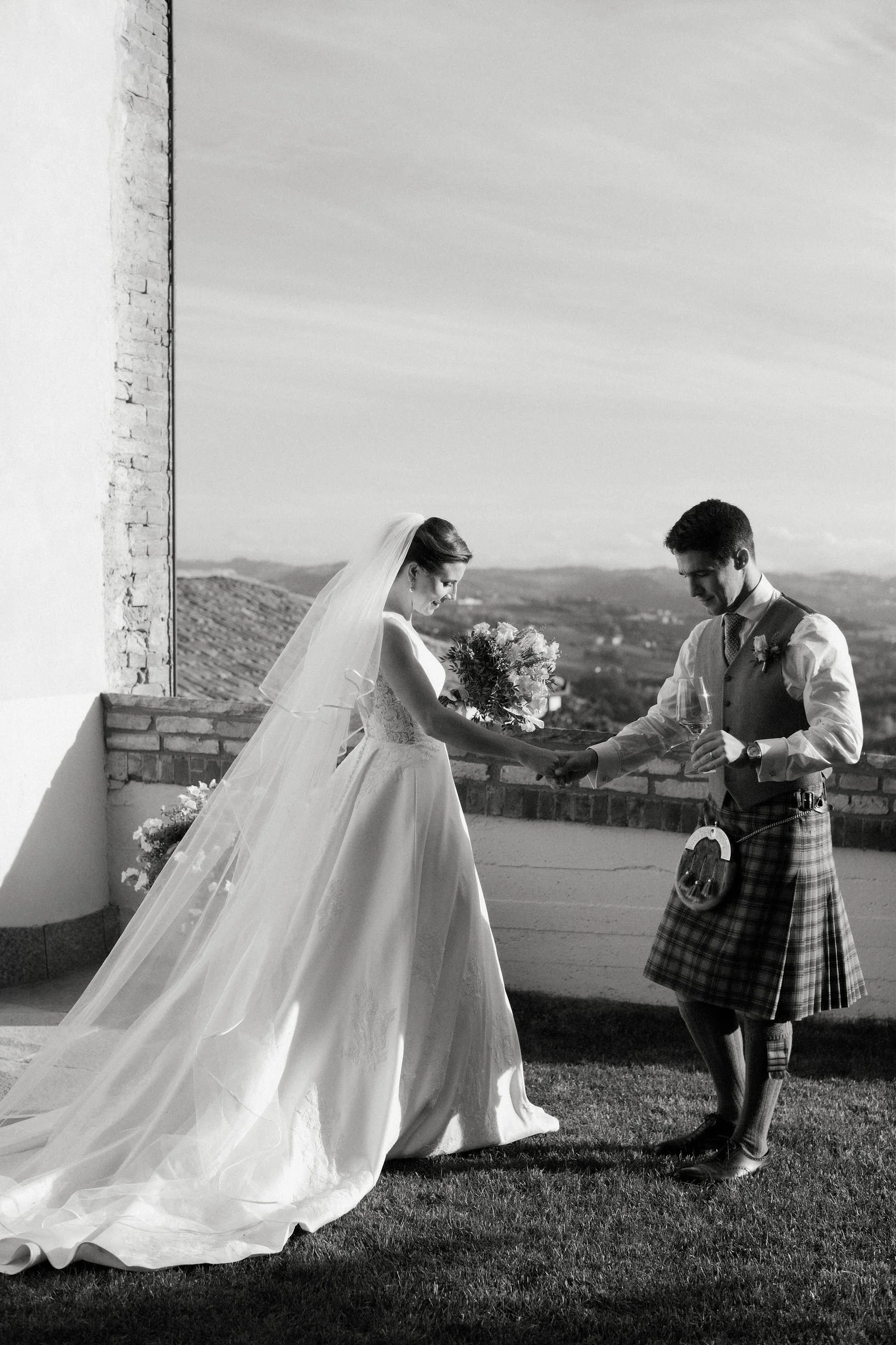 Una coppia di sposi si tiene per mano su un terrazzo all'aperto, lei indossa un abito da sposa con velo e lui indossa un kilt scozzese e una giacca, con un bicchiere di vino in mano.