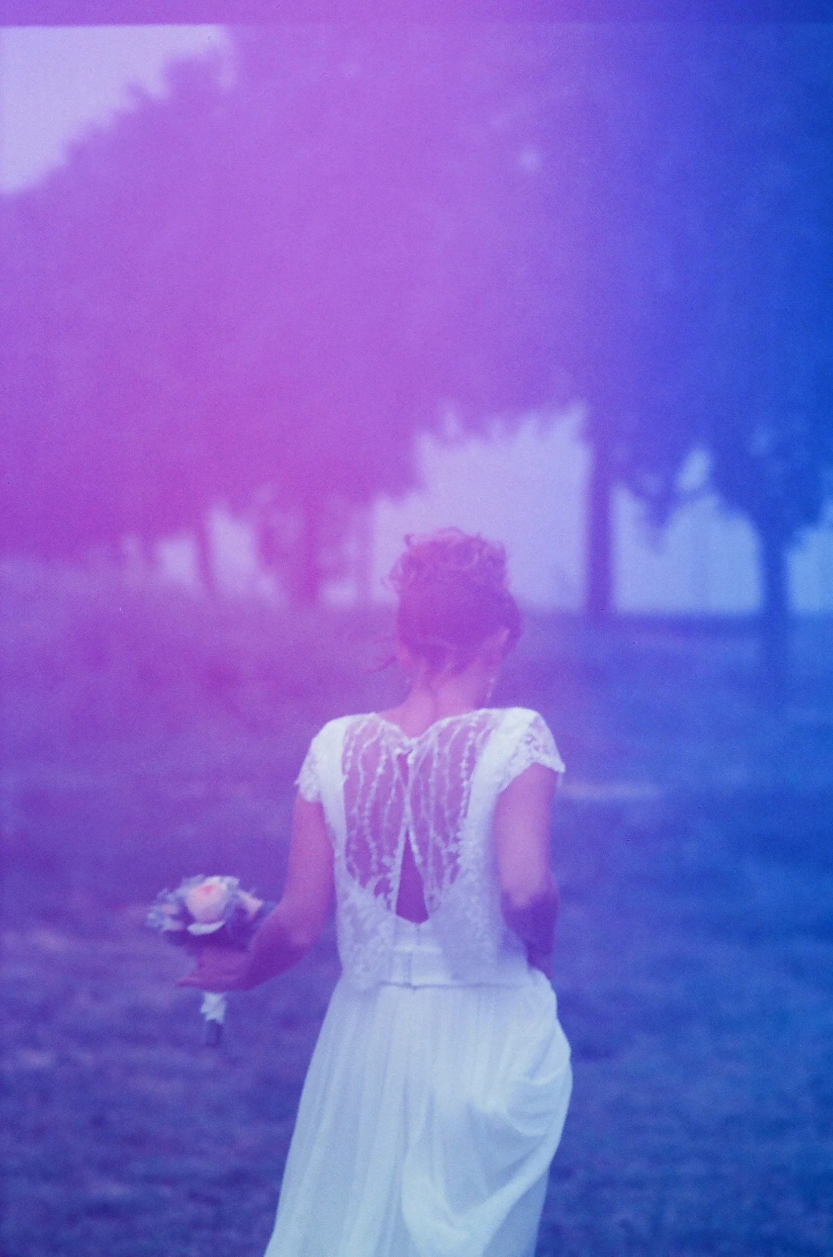 Una ragazza con capelli a riccio corti, indossa un vestito bianco lungo, tiene un mazzo di fiori e cammina in un parco con alberi al tramonto, con tonalità di colore blu e viola.