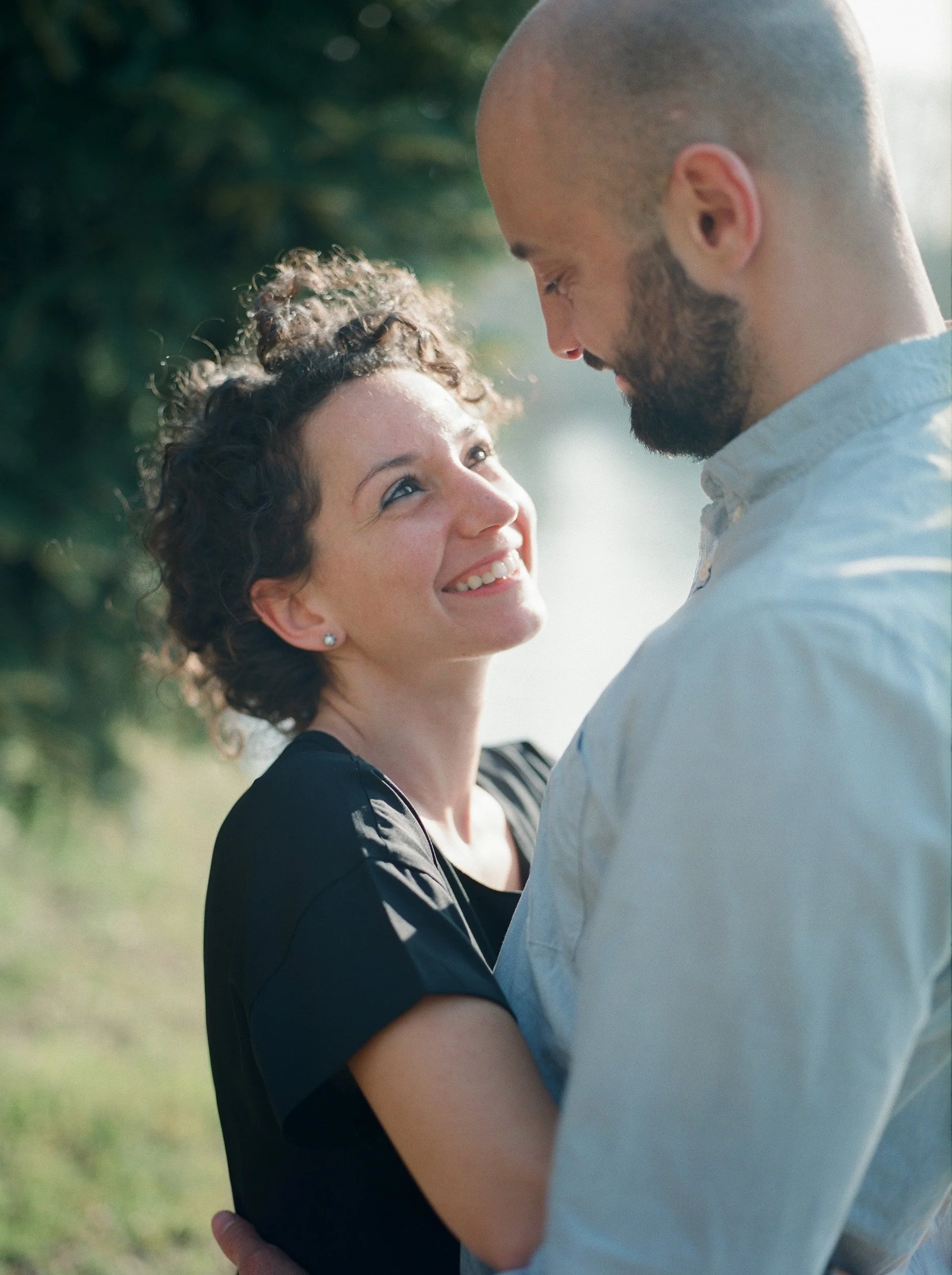 Una coppia si abbraccia all'aperto sotto la luce del sole con sfondo di alberi verdi, sorridendo e guardandosi negli occhi.