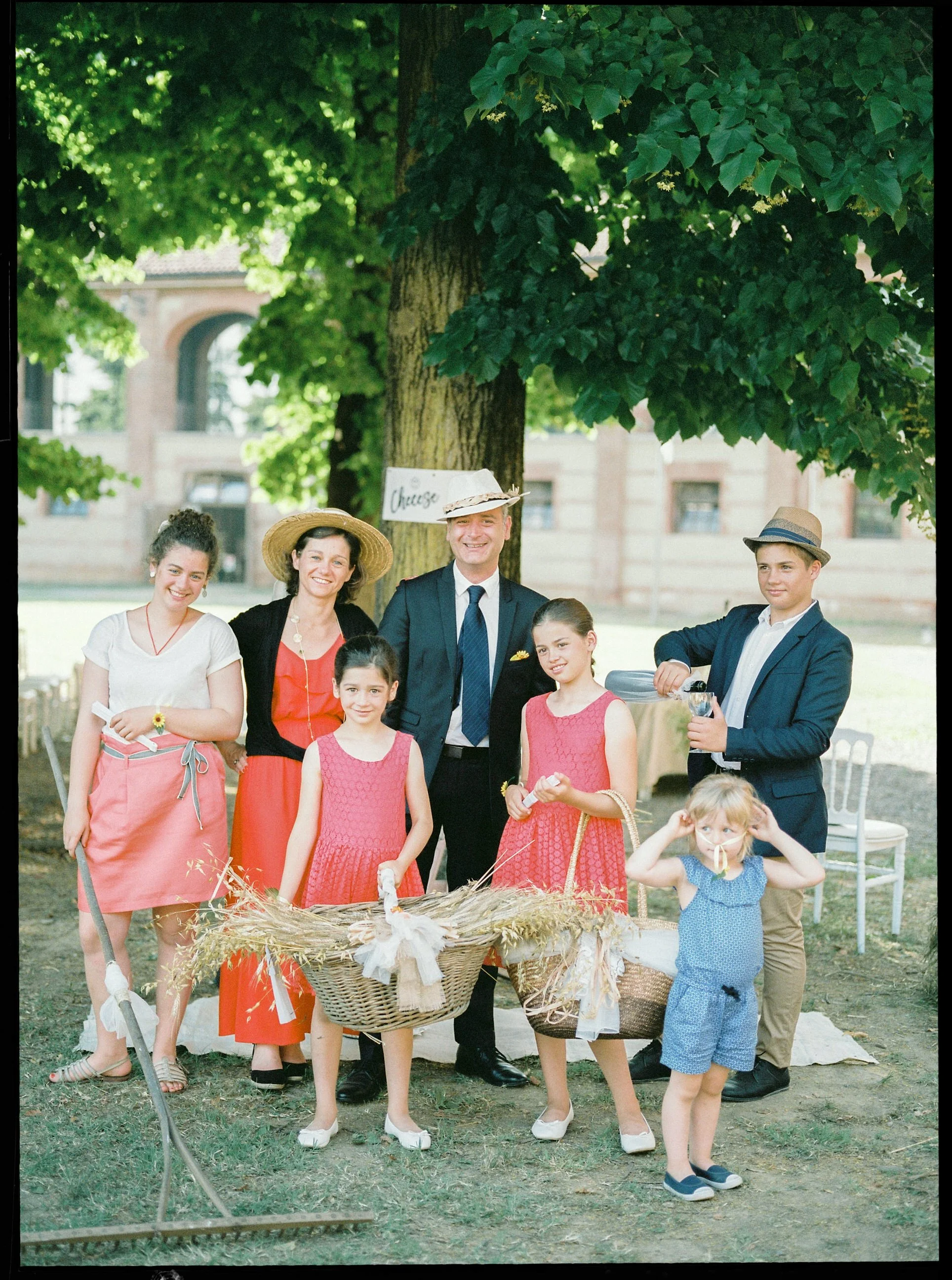 Un gruppo di persone, adulti e bambini, posano all'aperto sotto un albero durante un evento o celebrazione. Alcuni indossano cappelli e abiti colorati. C'è un cesto decorato con fiori e paglia al centro del gruppo.