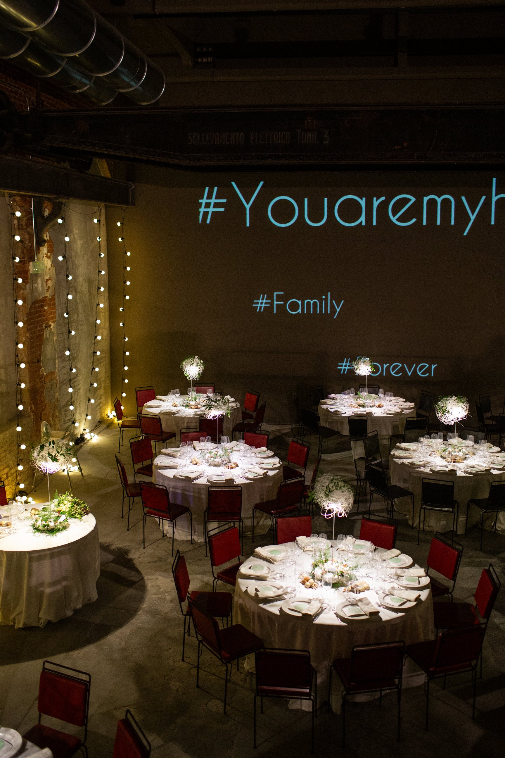 Sala elegante con tavoli rotondi decorati per un evento, luci soffuse e decorazioni floreali, con un grande schermo che mostra hashtag e parole in inglese, ambiente raffinato per occasioni speciali.