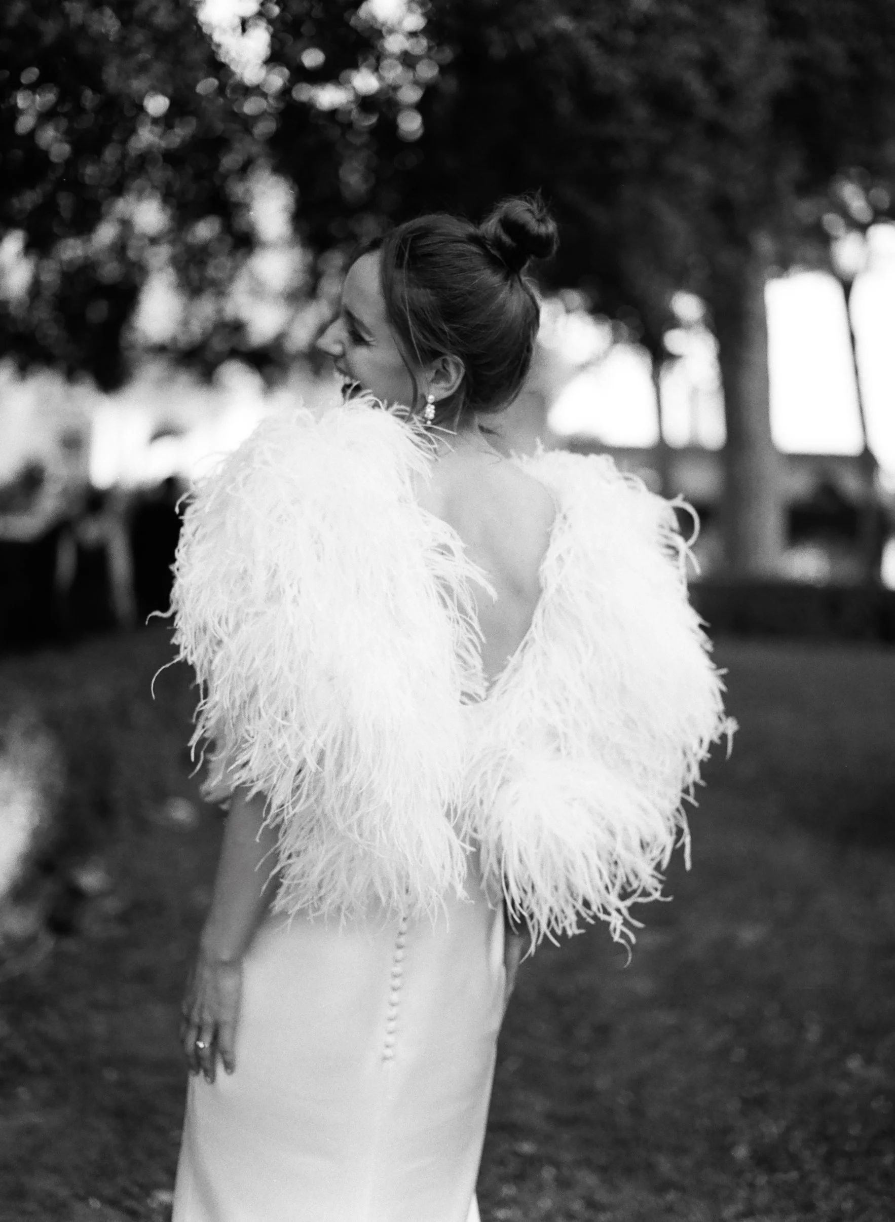 Una donna vestita con un abito lungo e una giacca con pelliccia bianca, che sorride e si gira di lato, in un ambiente all'aperto con alberi e luci sfocate sullo sfondo.