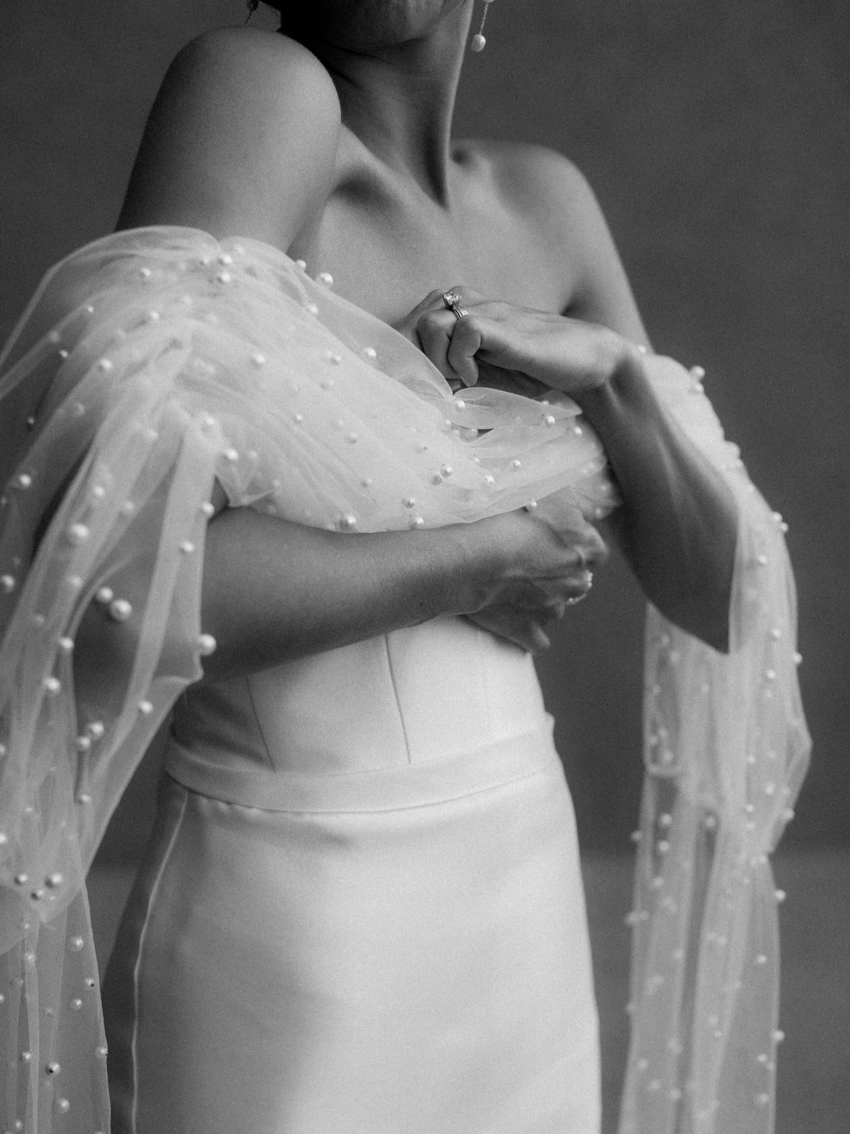 Dettaglio in bianco e nero di una donna con braccia incrociate, indossa un abito con maniche trasparenti decorato con perle e un anello di diamanti. Nella foto si evidenzia il collo, le spalle e una parte della mano.
