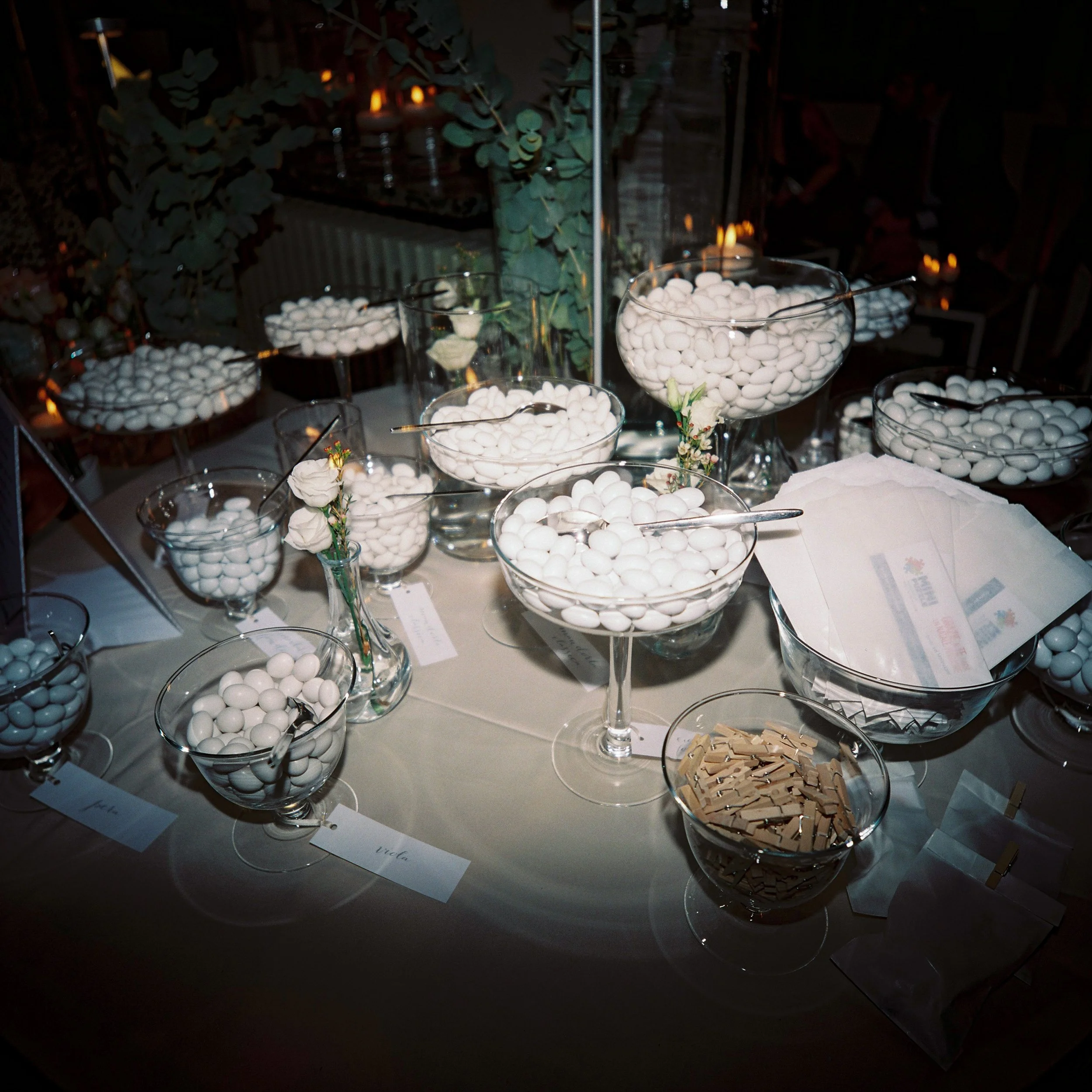 Tavolo decorato con numerosi vasi di cristallo trasparente pieni di palline bianche, alcune decorazioni floreali e colture. Sullo sfondo si vedono fumatori e luci calde, possibly a cena o un evento festivo.