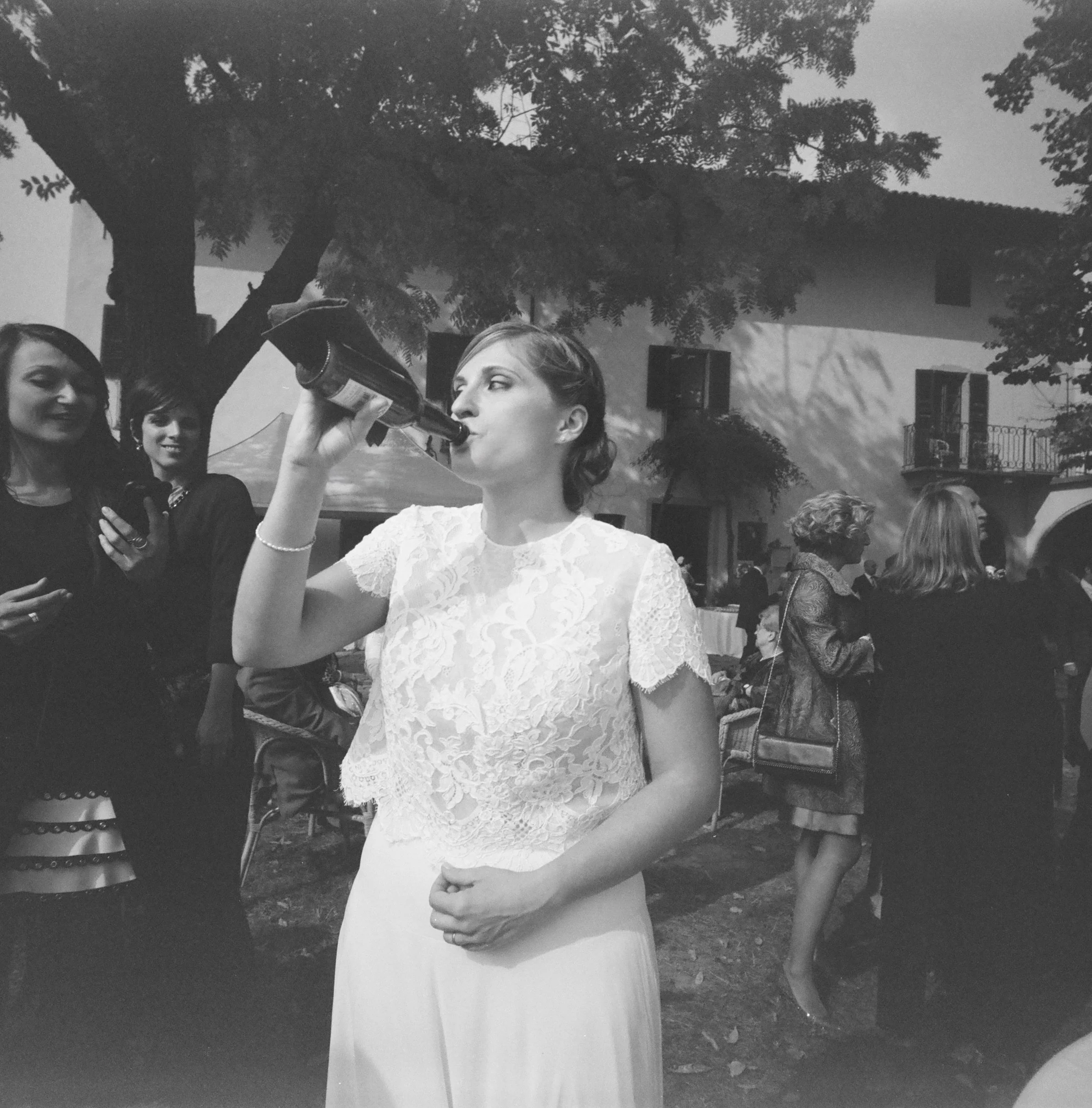 Ragazza con abito bianco scuro e capelli raccolti tiene in mano una bottiglia mentre sorride al fianco di altre persone in un evento all'aperto, con alberi e un edificio sullo sfondo.