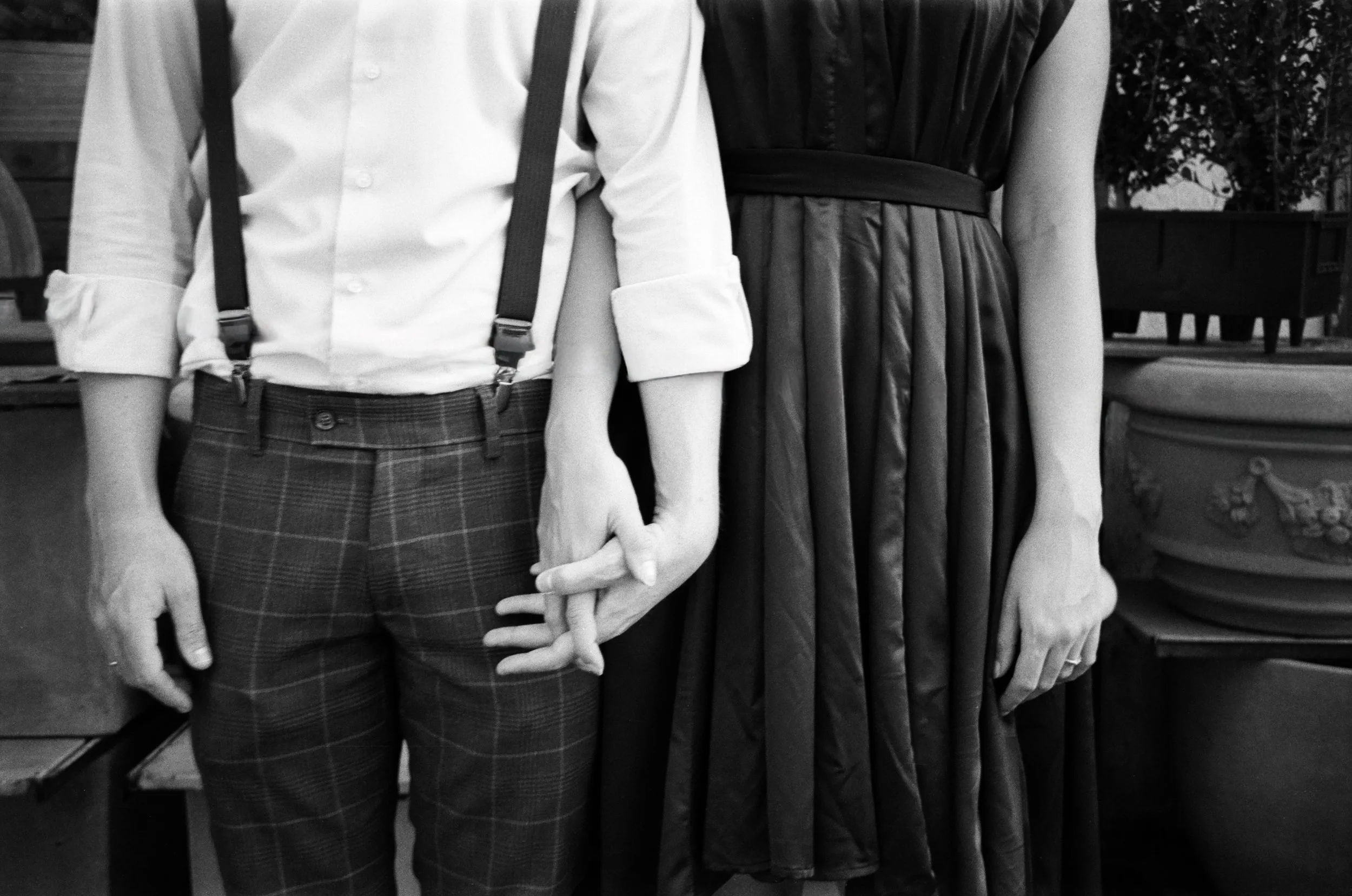 Due persone si tengono per mano, una con una camicia bianca a maniche lunghe e pantaloni a quadri, l'altra con un vestito scuro, in un ambiente interno con panchine in legno e piante sullo sfondo.