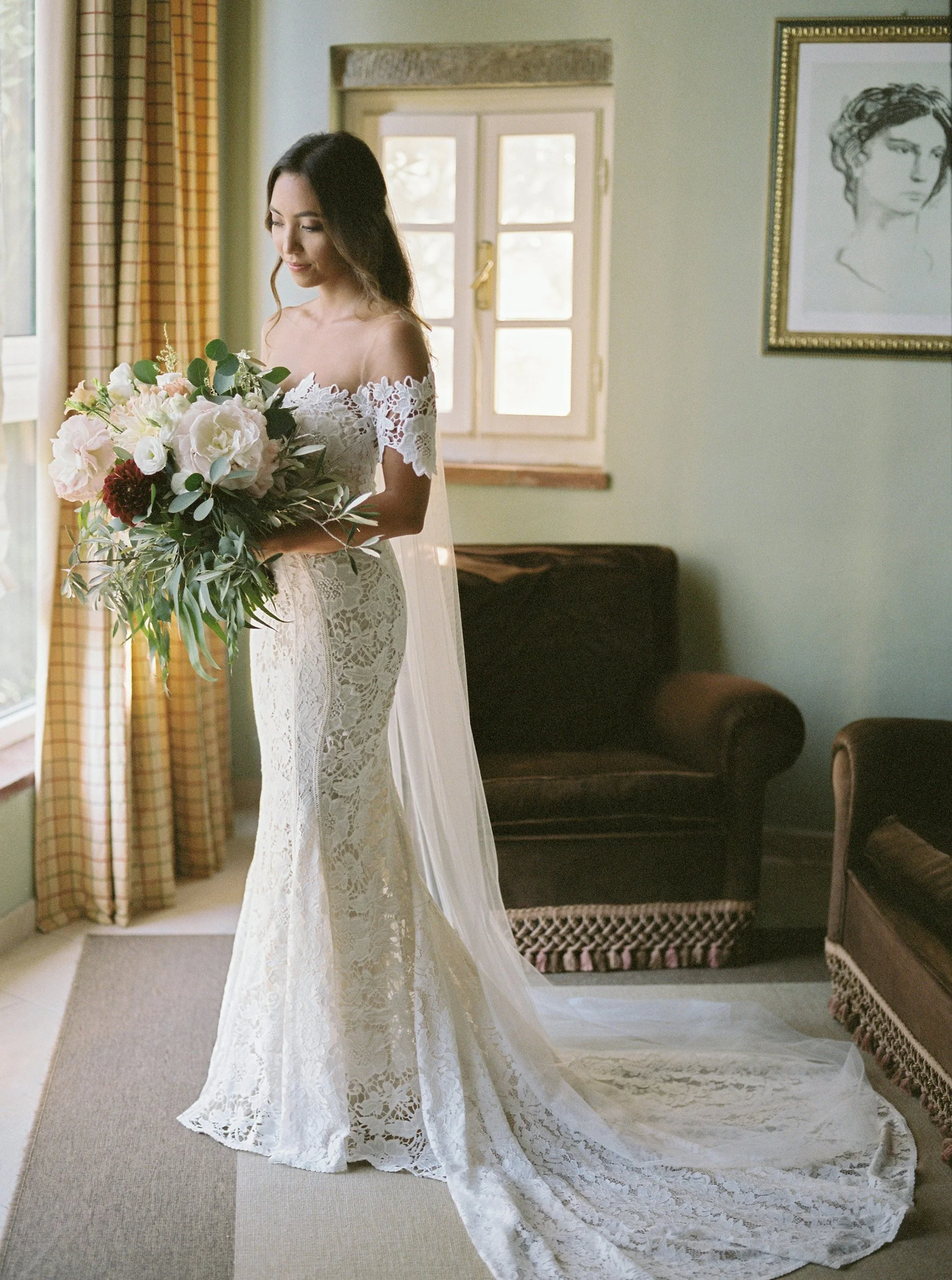 Donna vestita da sposa con abito bianco in pizzo, con corteggiatura e bouquet di fiori, in un soggiorno illuminato dalla luce naturale.
