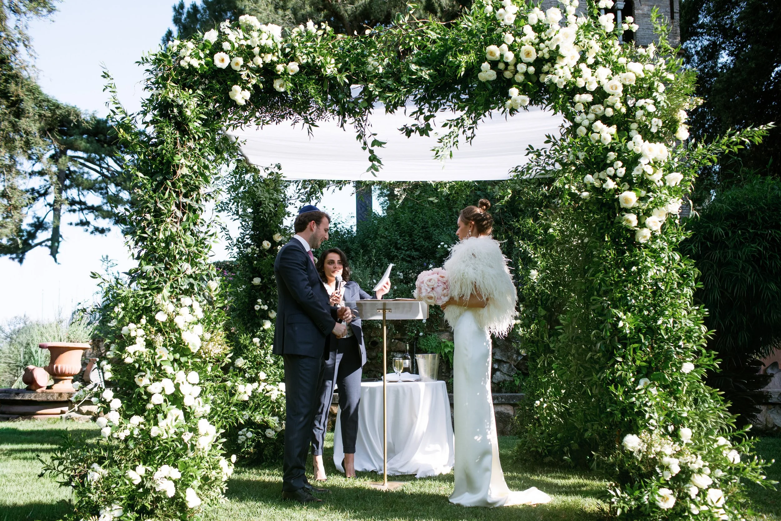 Coppia che si sposa sotto un arco decorato con fiori bianchi e foglie verdi, con una donna che tiene un microfono e un tappeto bianco sopra un tavolo, in un giardino all'aperto.