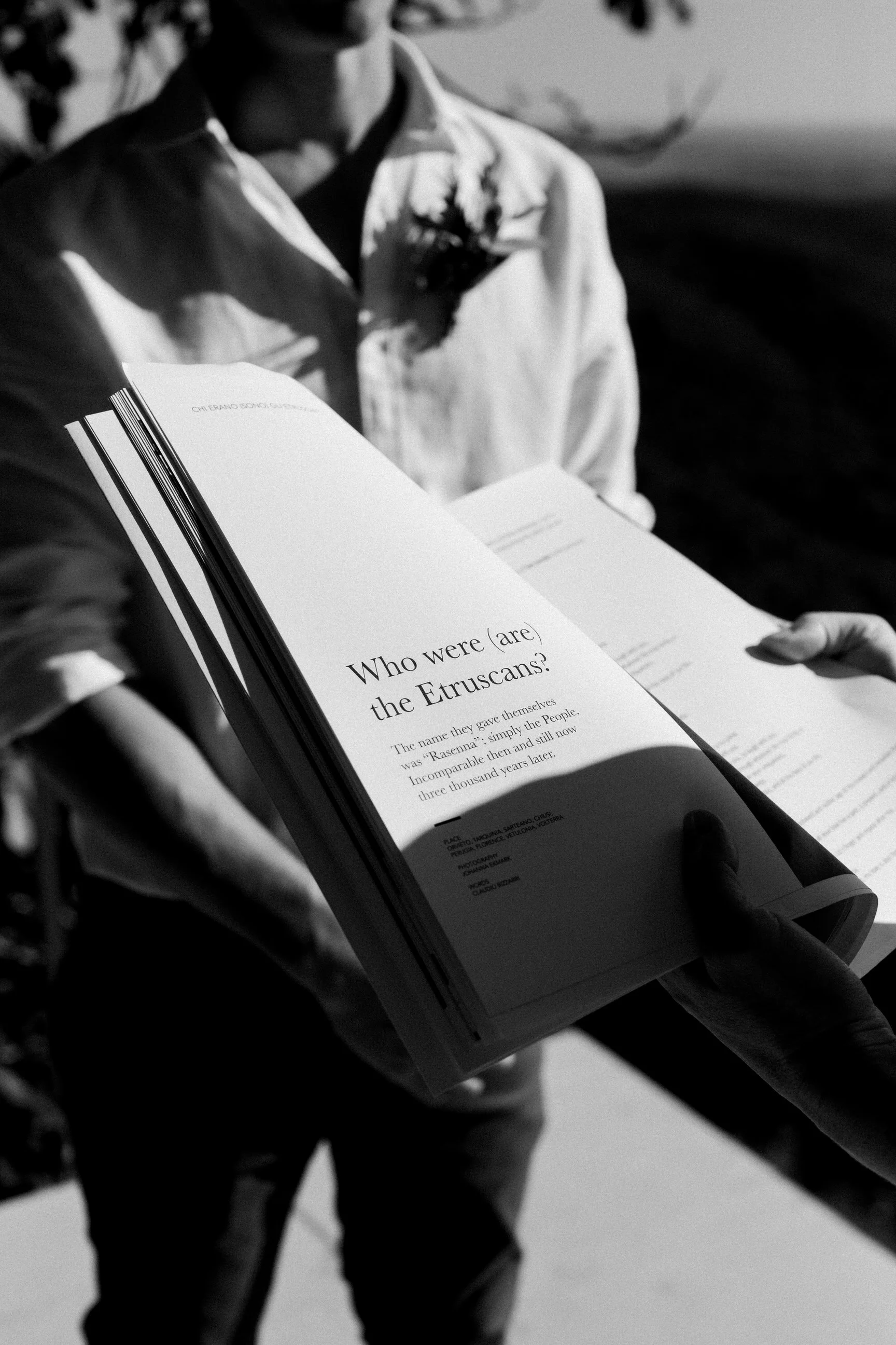 Una persona tiene in mano un libro aperto con un testo in inglese e italiano, visibile una domanda sul passato dei Etruschi, mentre indossa una camicia bianca con un disegno scuro. La scena è in bianco e nero.