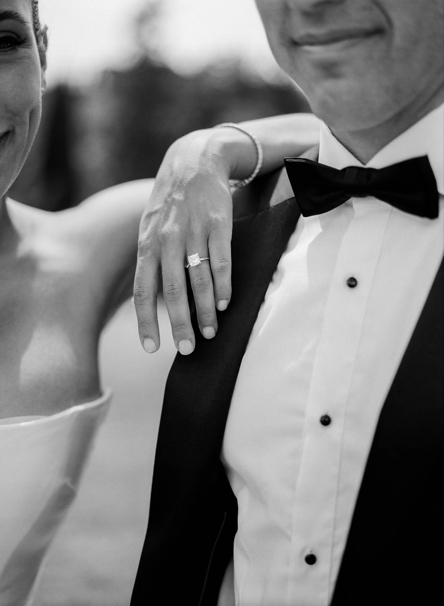 Dettaglio di una coppia in abiti formali, con attenzione sulla mano della donna che indossa un anello di diamante, e sulla spalla del uomo che indossa un abito con papillon.