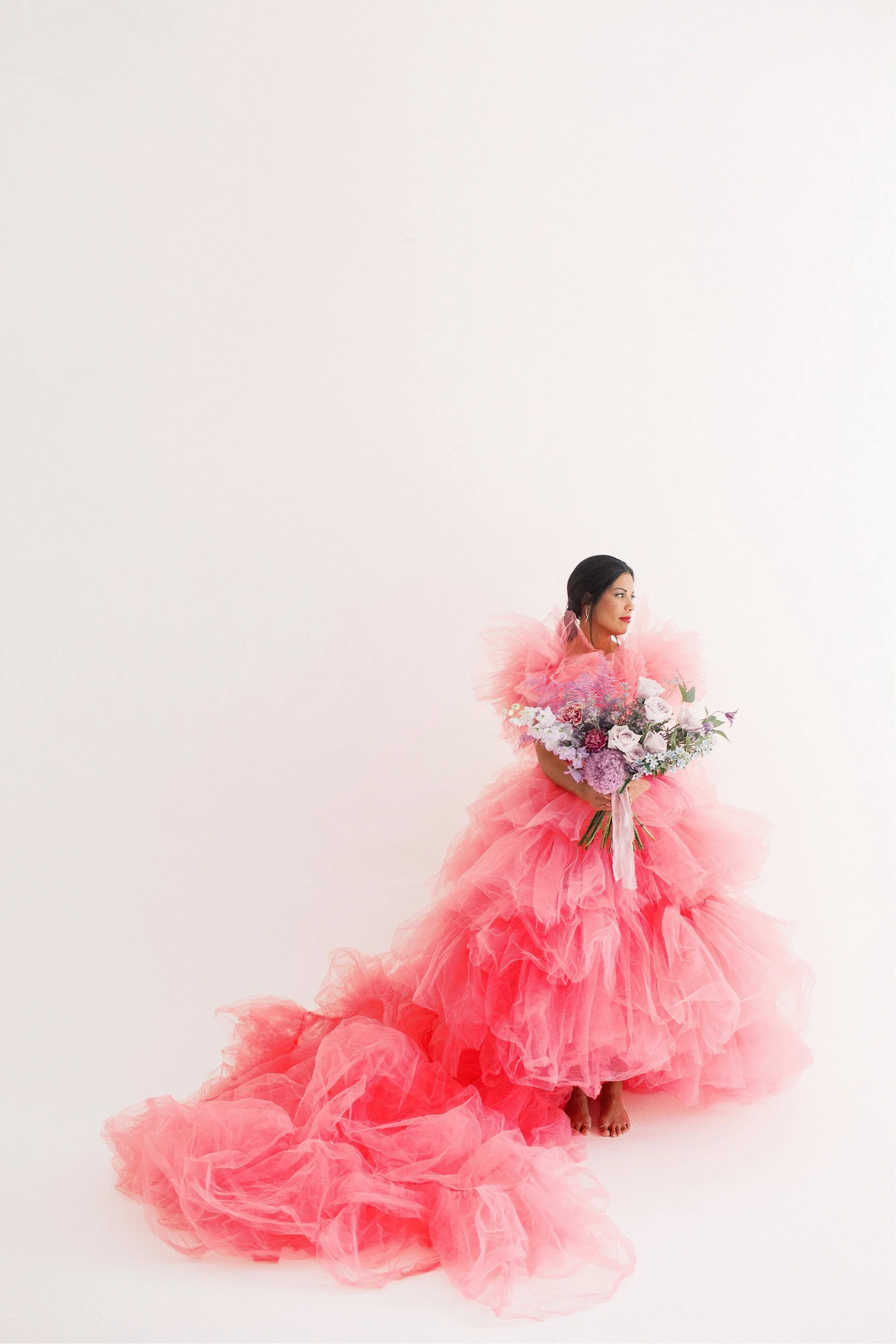 Una donna vestita con un abito rosa a strati di tulle tiene un mazzo di fiori multicolori, si trova su uno sfondo bianco.