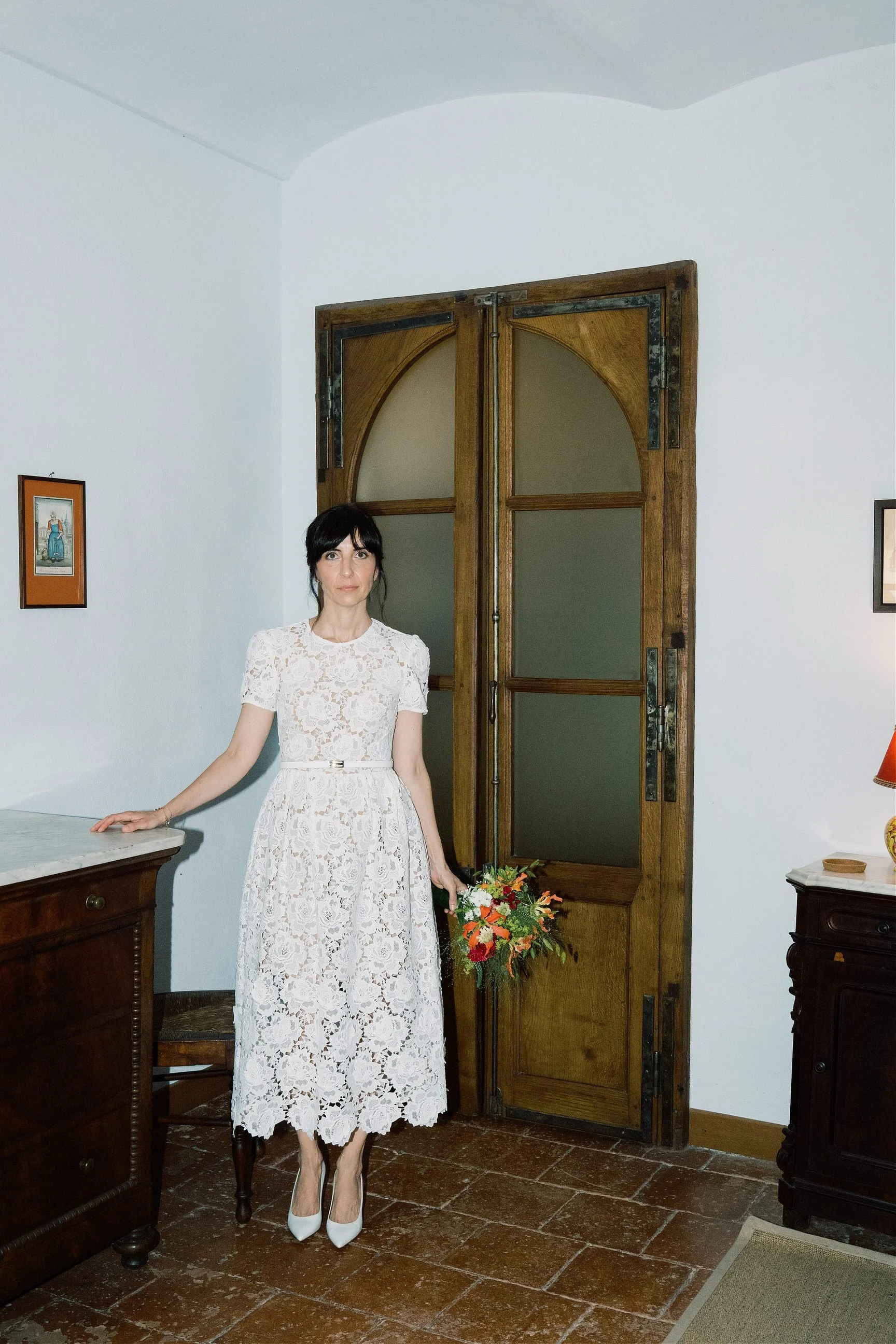 Una donna con vestito lungo bianco con inserti in pizzo, scarpe bianche, porta un bouquet di fiori e si trova in una stanza con mobili in legno e pareti bianche.