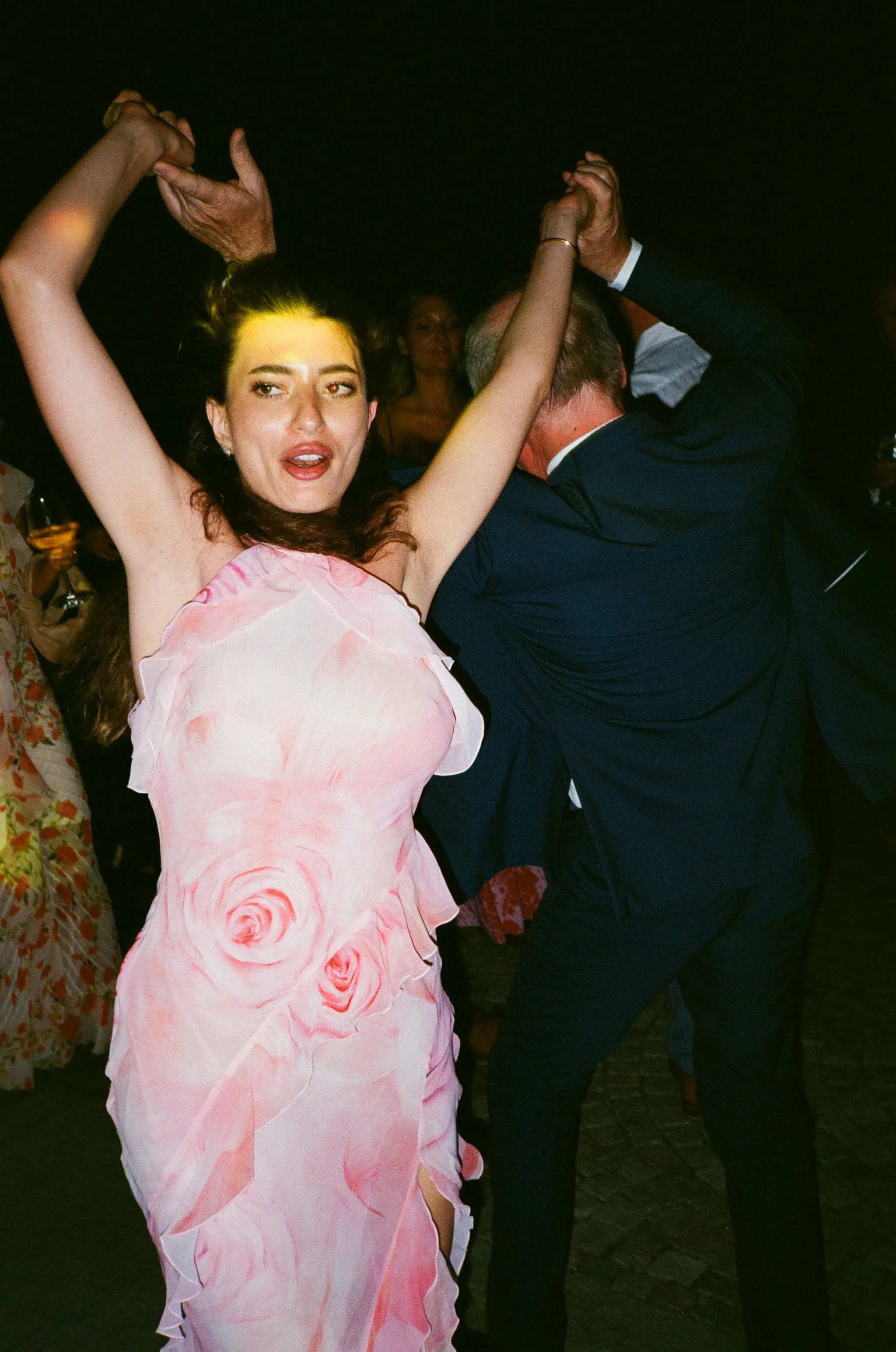 Una coppia di adulti che balla, vista di lato, con una donna in primo piano che indossa un vestito rosa con motivo floreale e un uomo in abito nero.