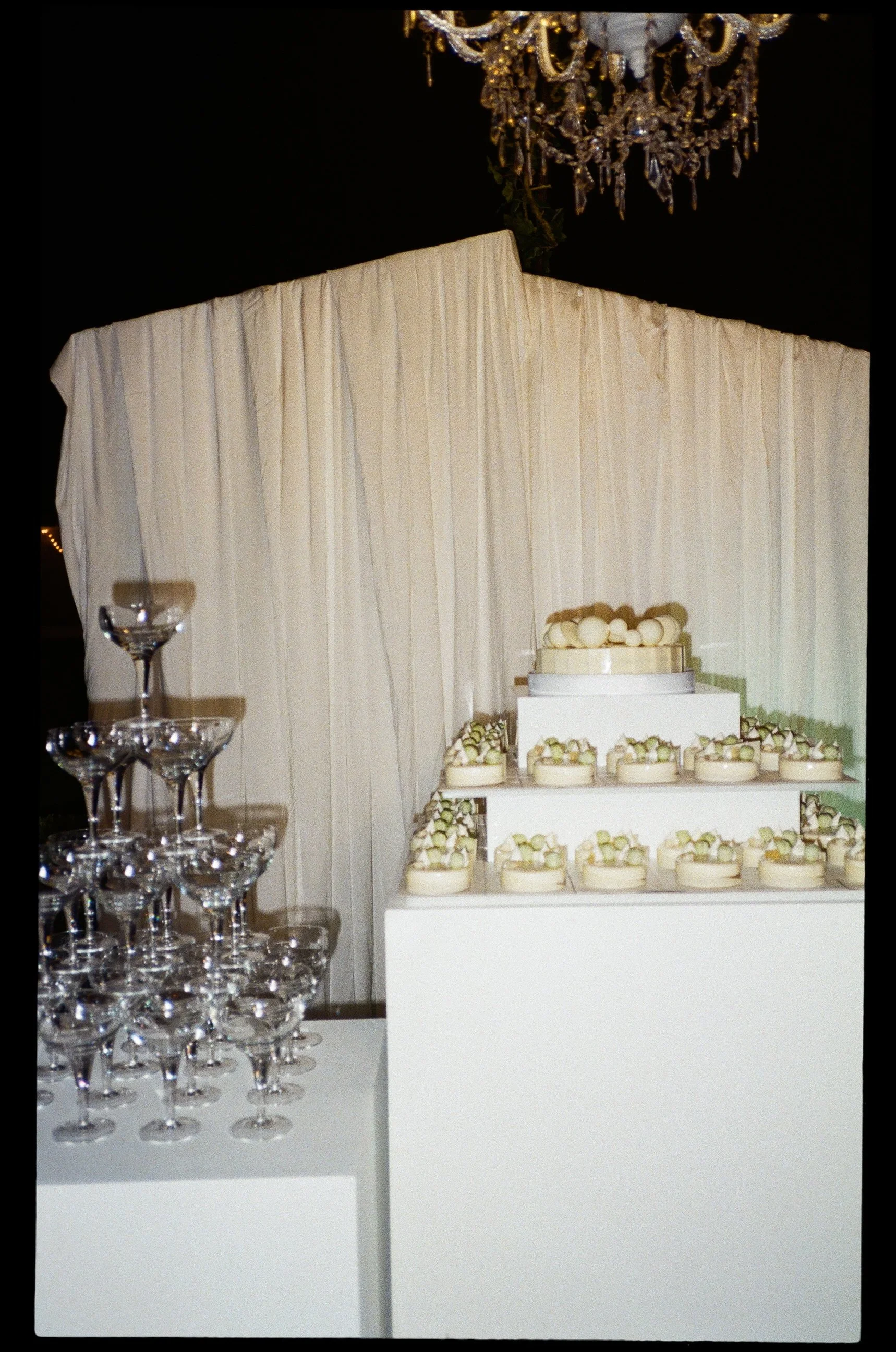 Tavolo con cioccolatini decorativi e bicchieri di champagne in ambiente elegante, con drappeggi bianchi sullo sfondo e lampadario di cristallo sopra.