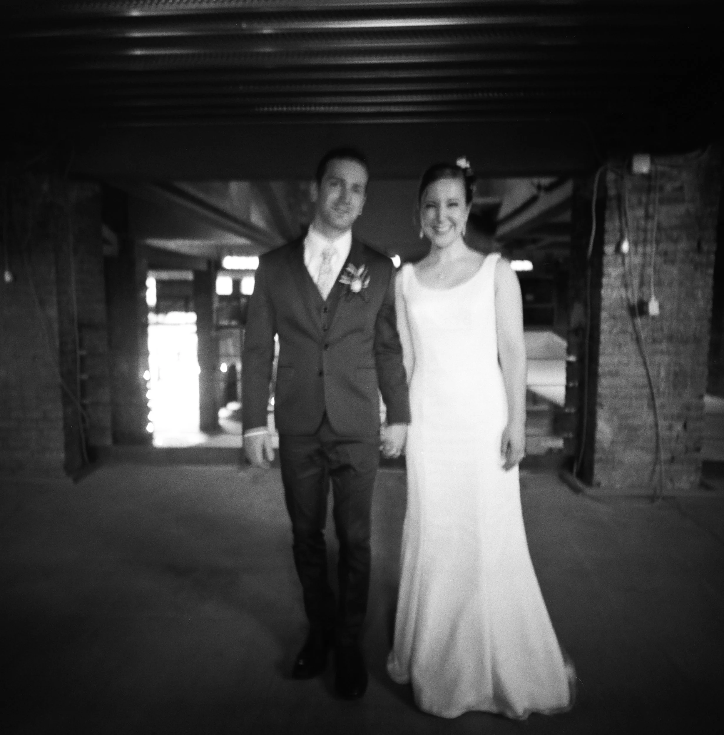 Foto in bianco e nero di una coppia sorridente vestita formalmente, sulla scena davanti a una parete di fusti di legno, in un ambiente interno, probabilmente una scena di matrimonio.