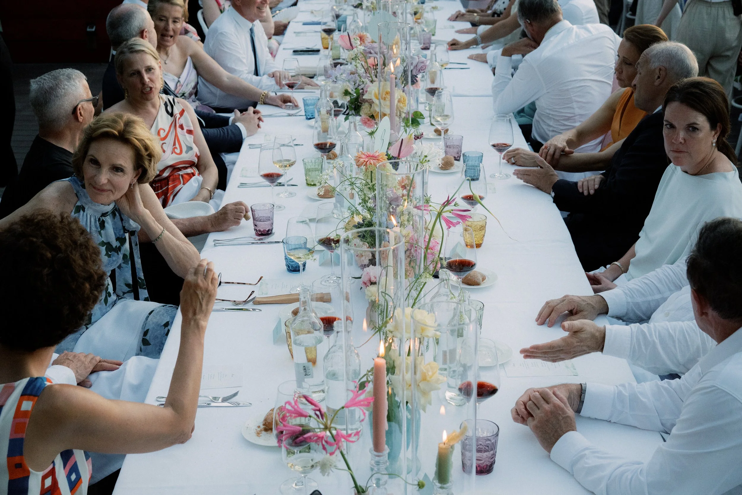 Una lunga tavola imbandita con fiori colorati e candele accese, circondata da persone sedute che partecipano a un pranzo o una festa elegante.