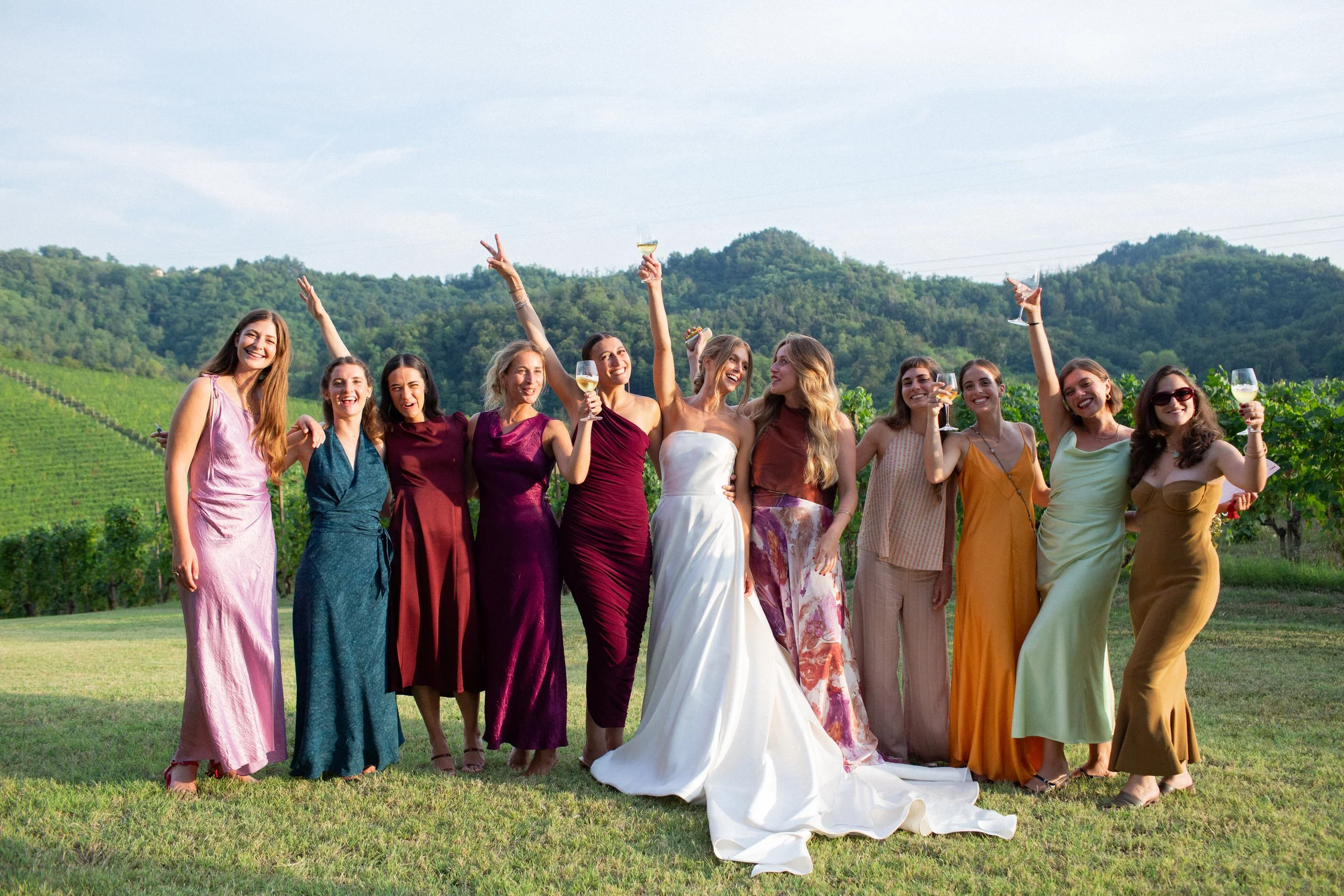 Un gruppo di donne festeggiano all'aperto in un campo con sfondo di colline verdi, tutte vestite con abiti colorati vistosi e alcune tengono bicchieri di vino.