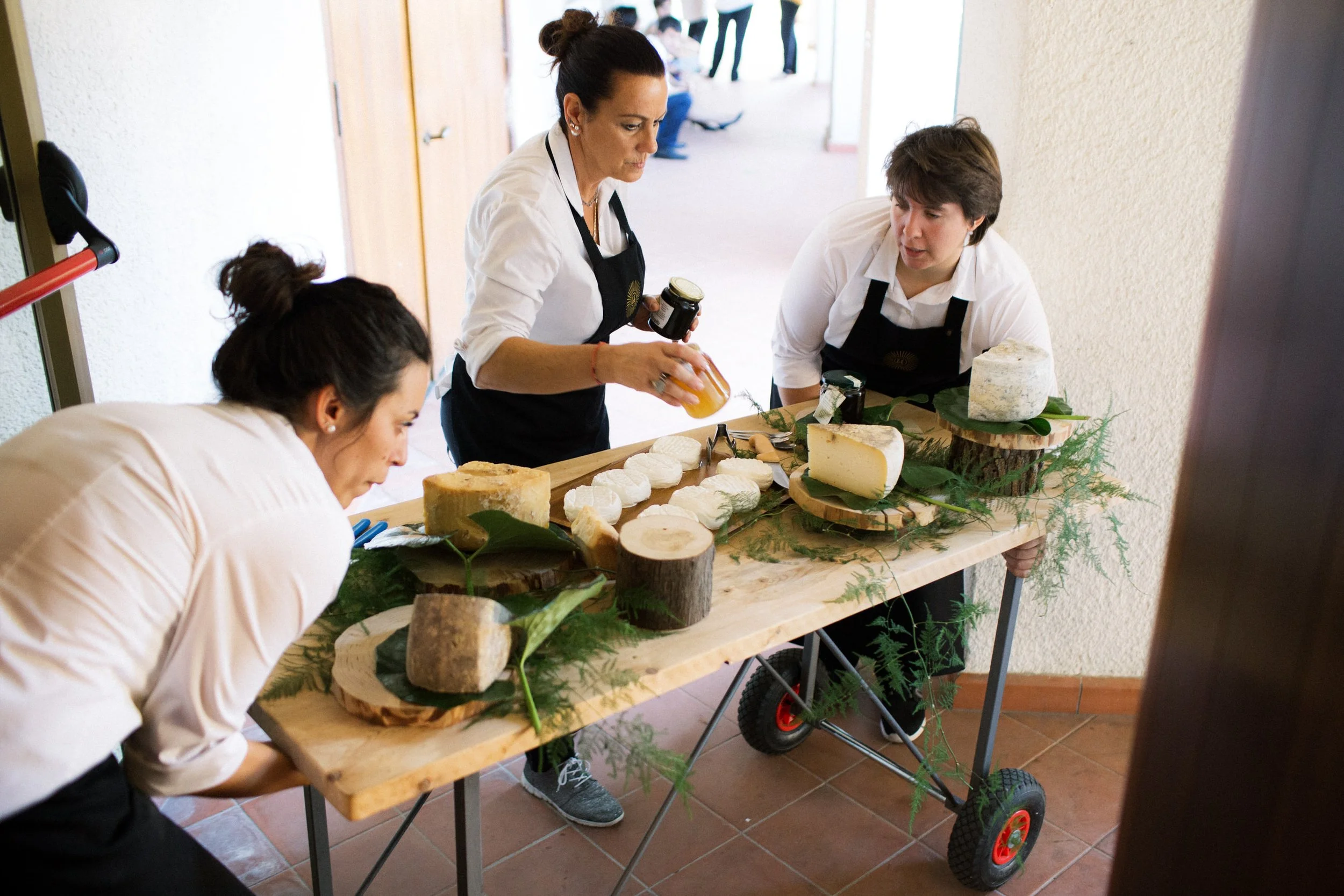 Tre donne che partecipano a una degustazione di formaggio su un tavolo di legno decorato con foglie e rami, con formaggi di diverse forme e dimensioni. Una donna, al centro, tiene una confezione di marmellata, mentre le altre sono curve di formaggio.