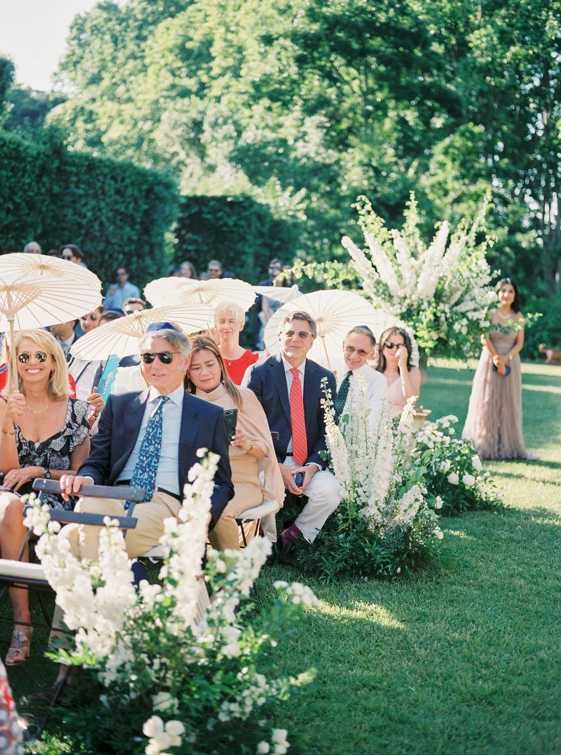 Gente seduta all'aperto sotto un cielo soleggiato, circondata da alberi verdi e decorazioni floreali, probabilmente a un matrimonio o evento sociale.