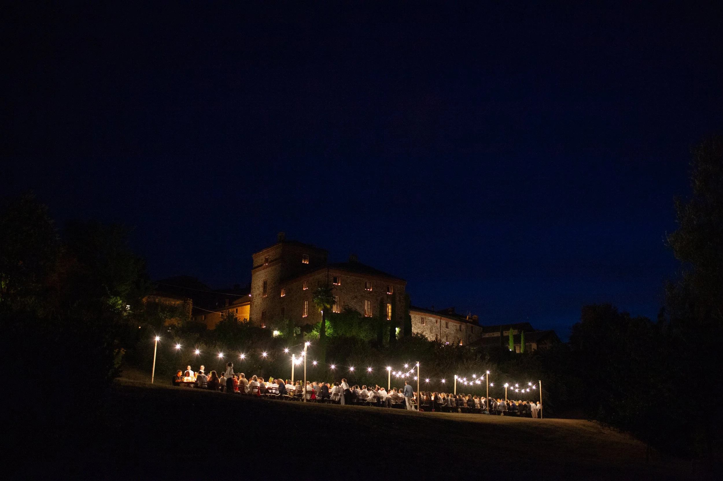 Gruppo di persone sedute a tavoli all'aperto sotto luci durante la sera, con un castello in pietra sullo sfondo e un cielo notturno.